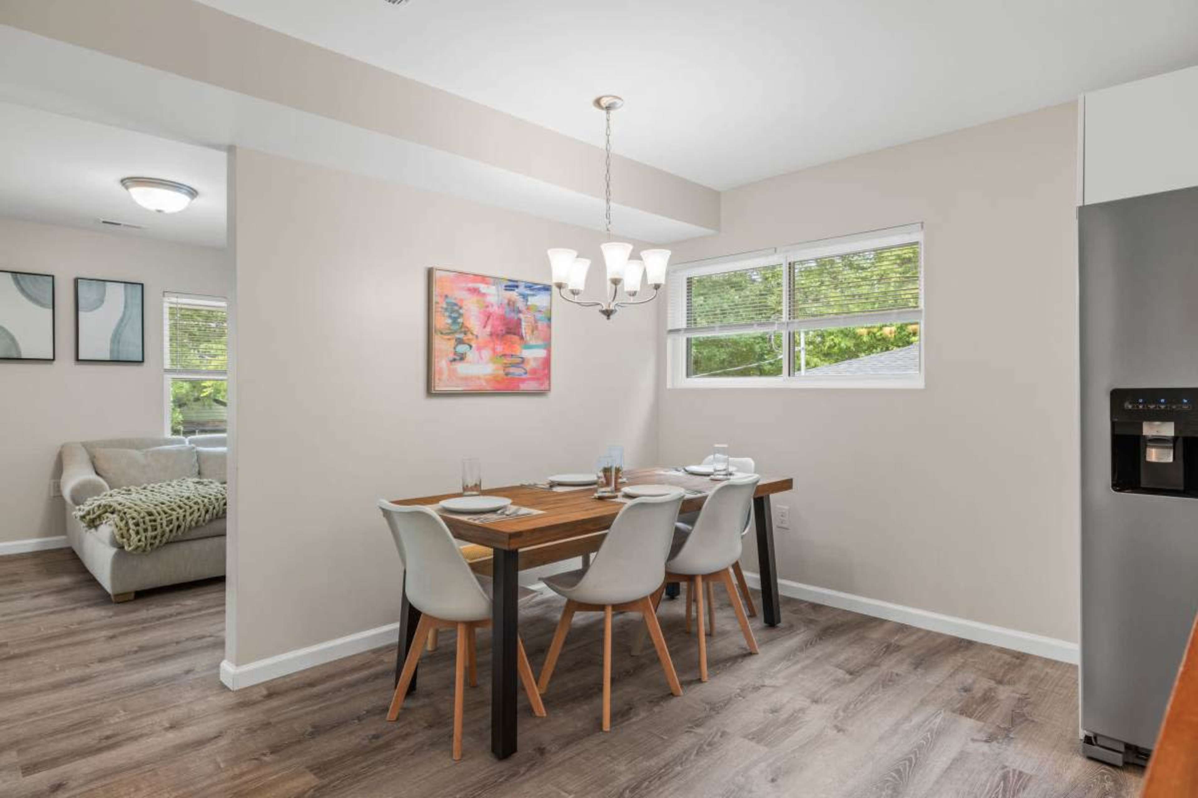 A dining area features a wooden table set for four, with a colorful artwork on the wall and a view of a living space in the background.