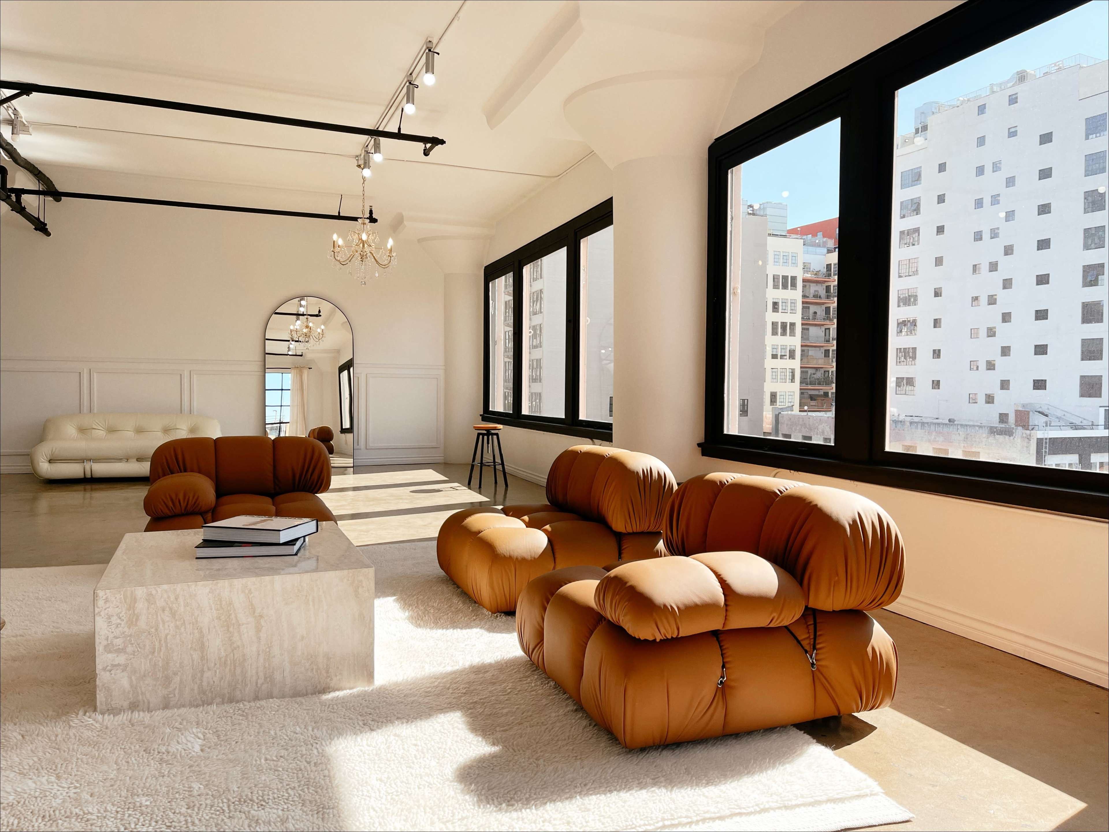 The image shows a spacious room with large windows, featuring brown, puffy sofas arranged around a marble coffee table on a light-colored rug.