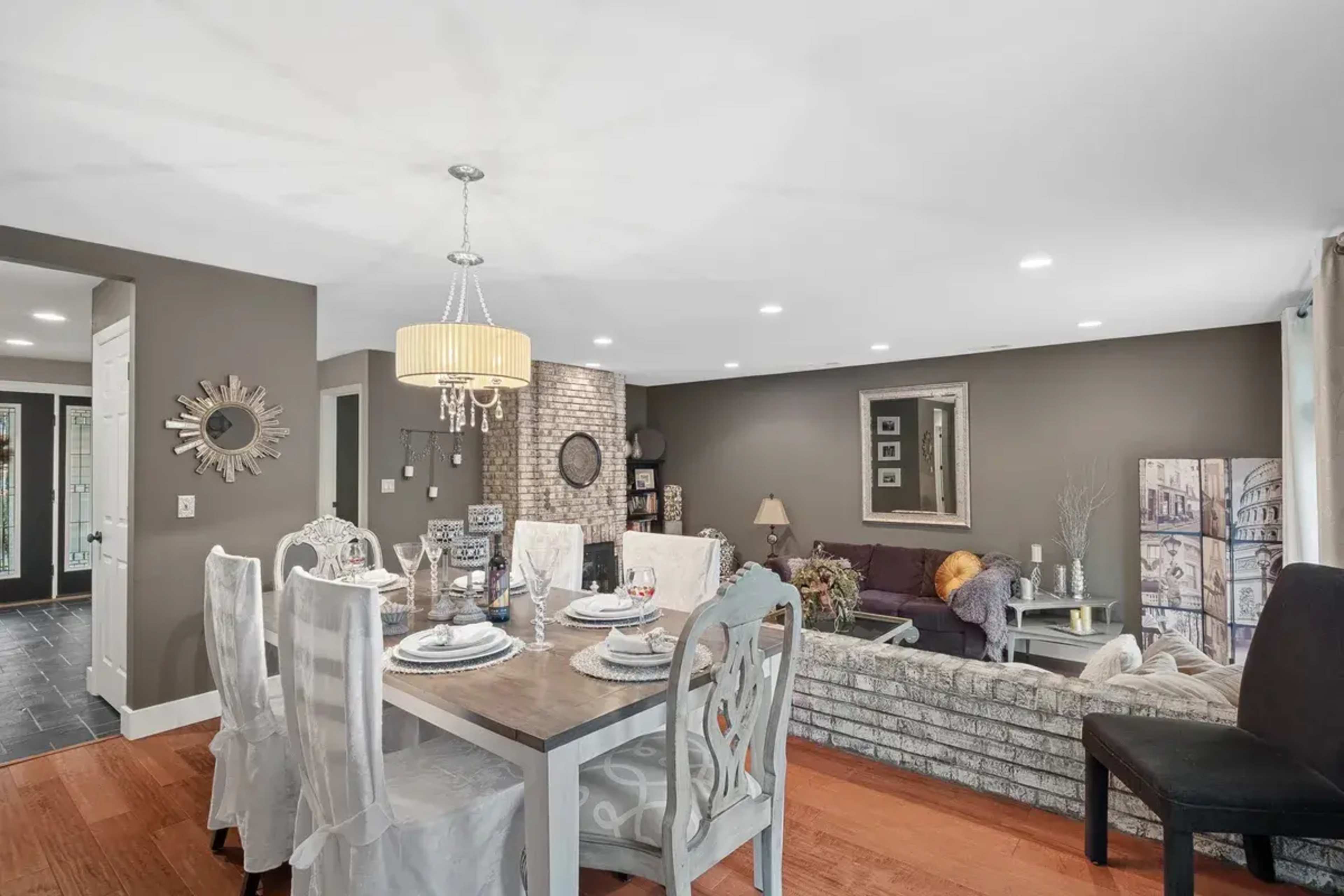 A well-lit dining area featuring a wooden table set for dinner, surrounded by white chairs, with a living room in the background that includes a gray sofa and decorative wall art.