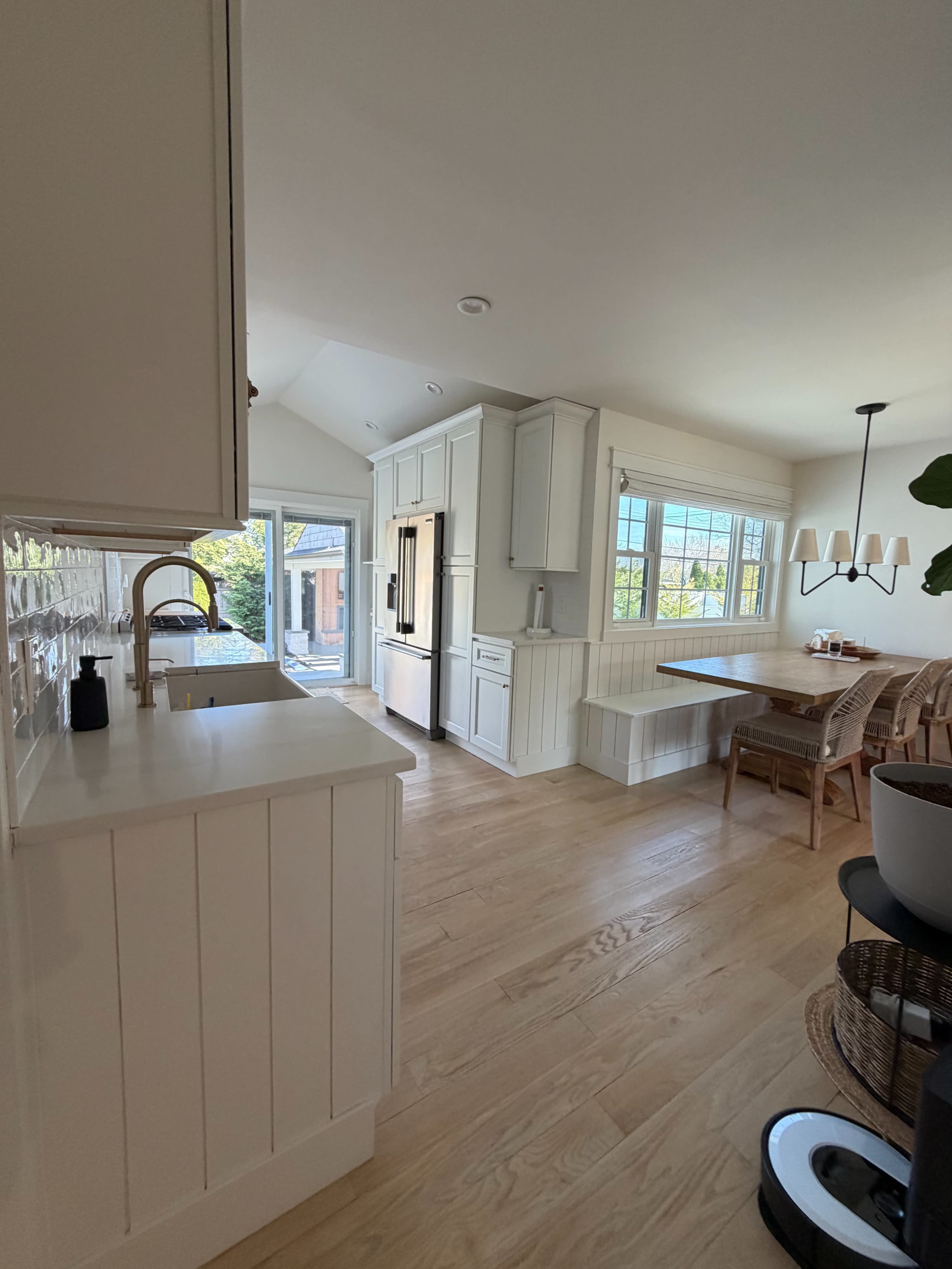The image shows a modern kitchen with white cabinetry, a refrigerator, a dining area with a wooden table, and large windows allowing natural light.