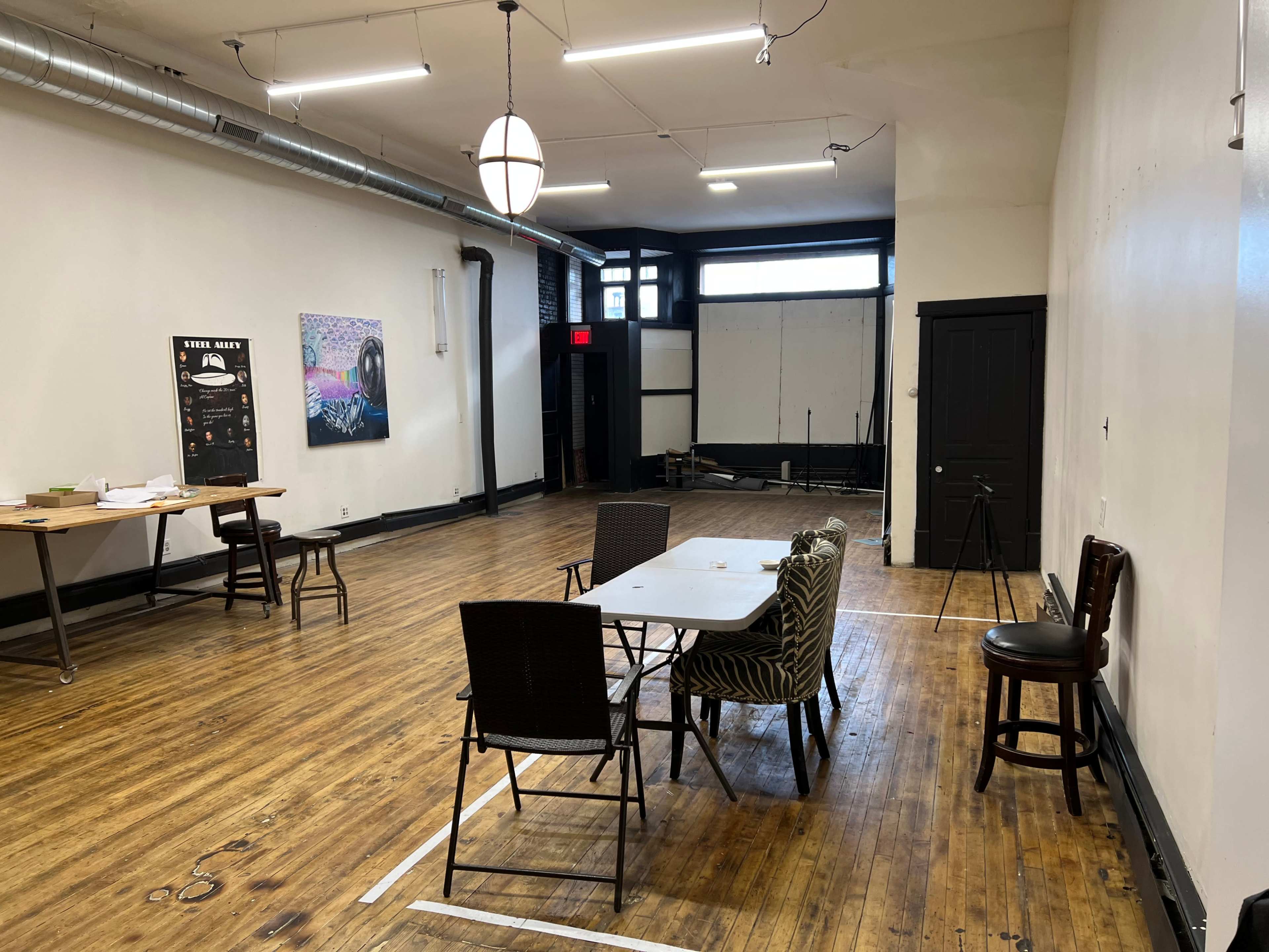 A spacious, empty room with wooden flooring, a few tables and chairs arranged, and large windows along one wall.