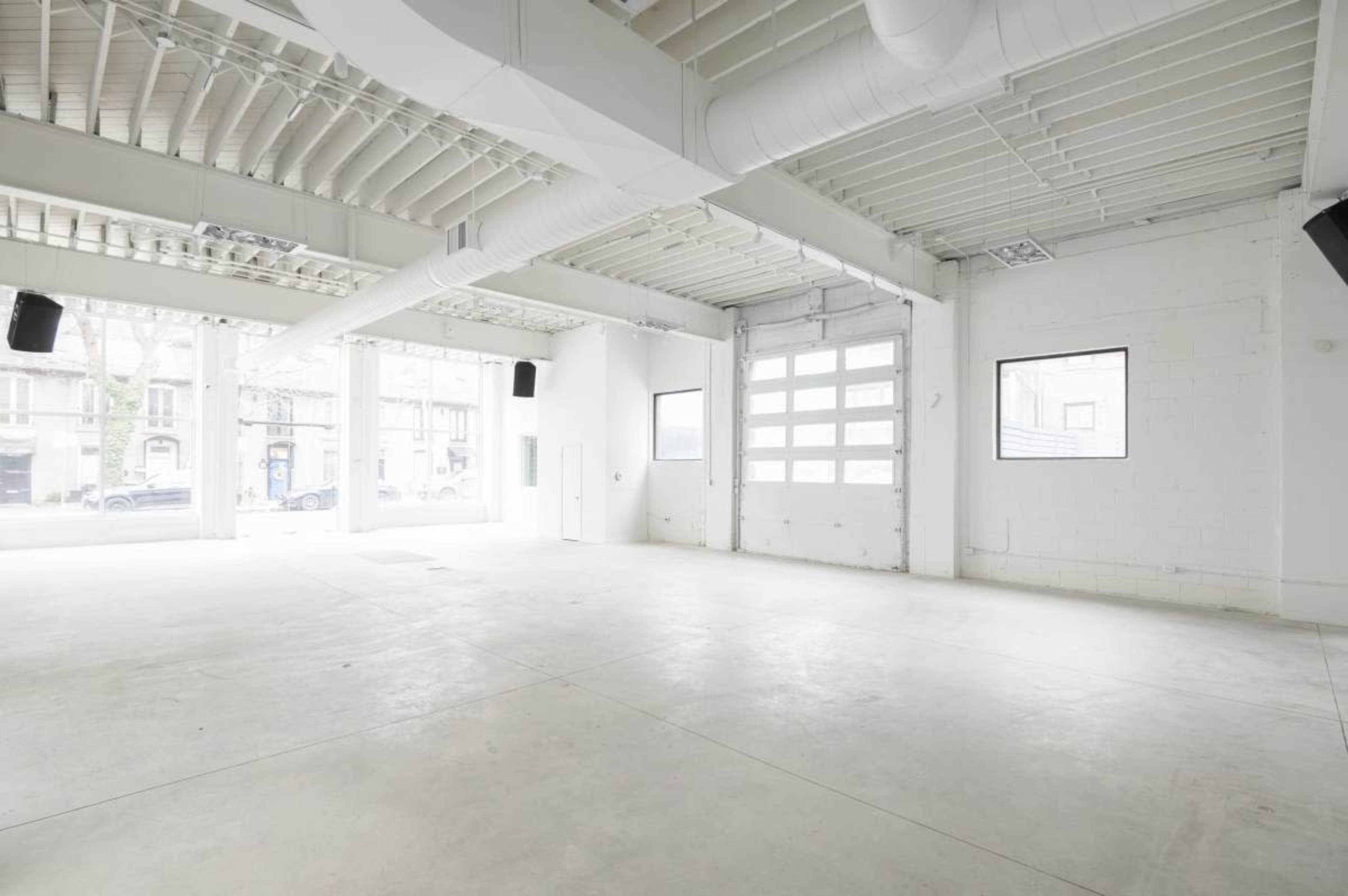 The image shows an empty, well-lit interior space with high ceilings, large windows, and a concrete floor.
