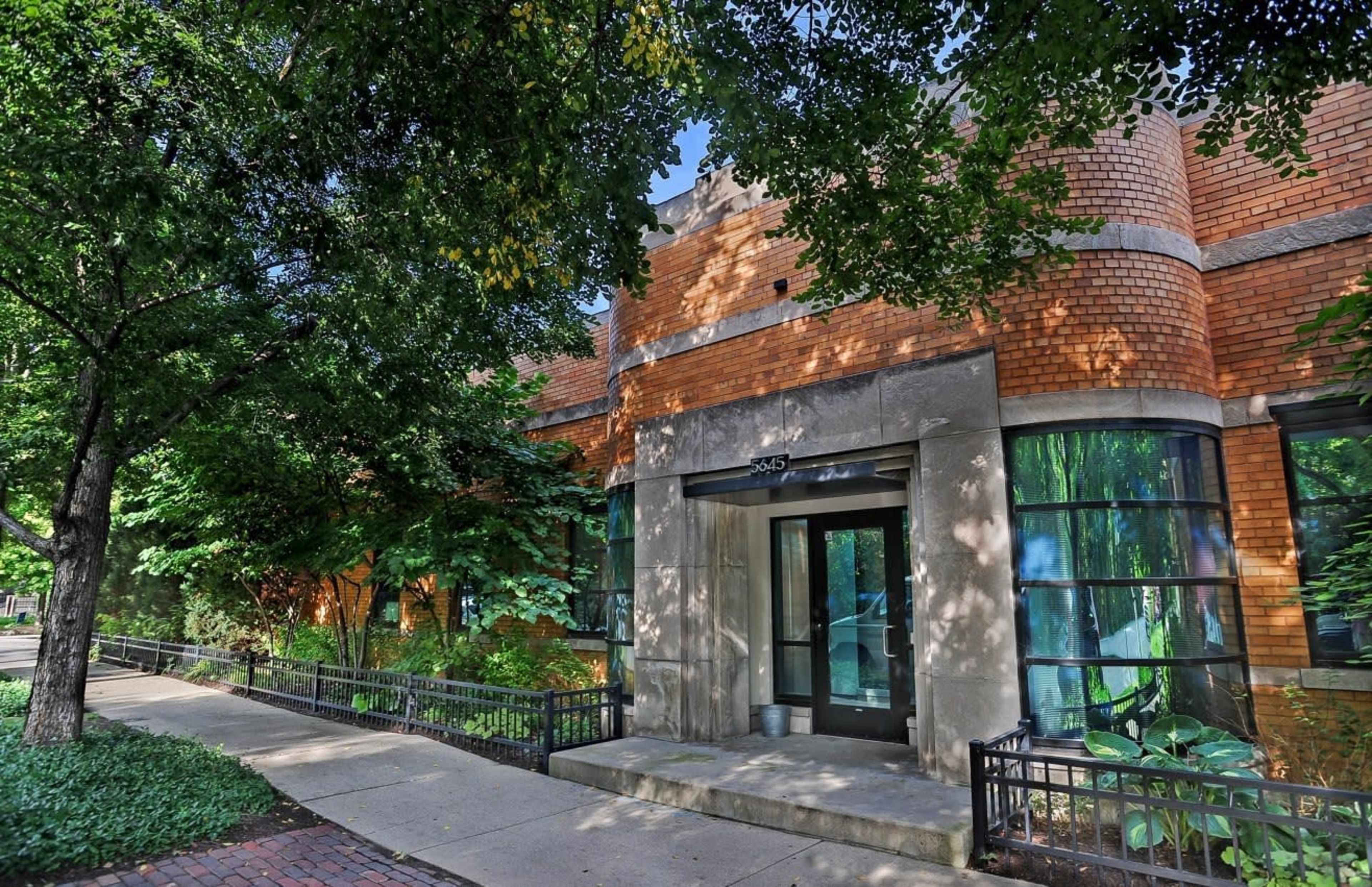 A brick building with large windows and a central entrance, surrounded by trees and greenery along a paved walkway.