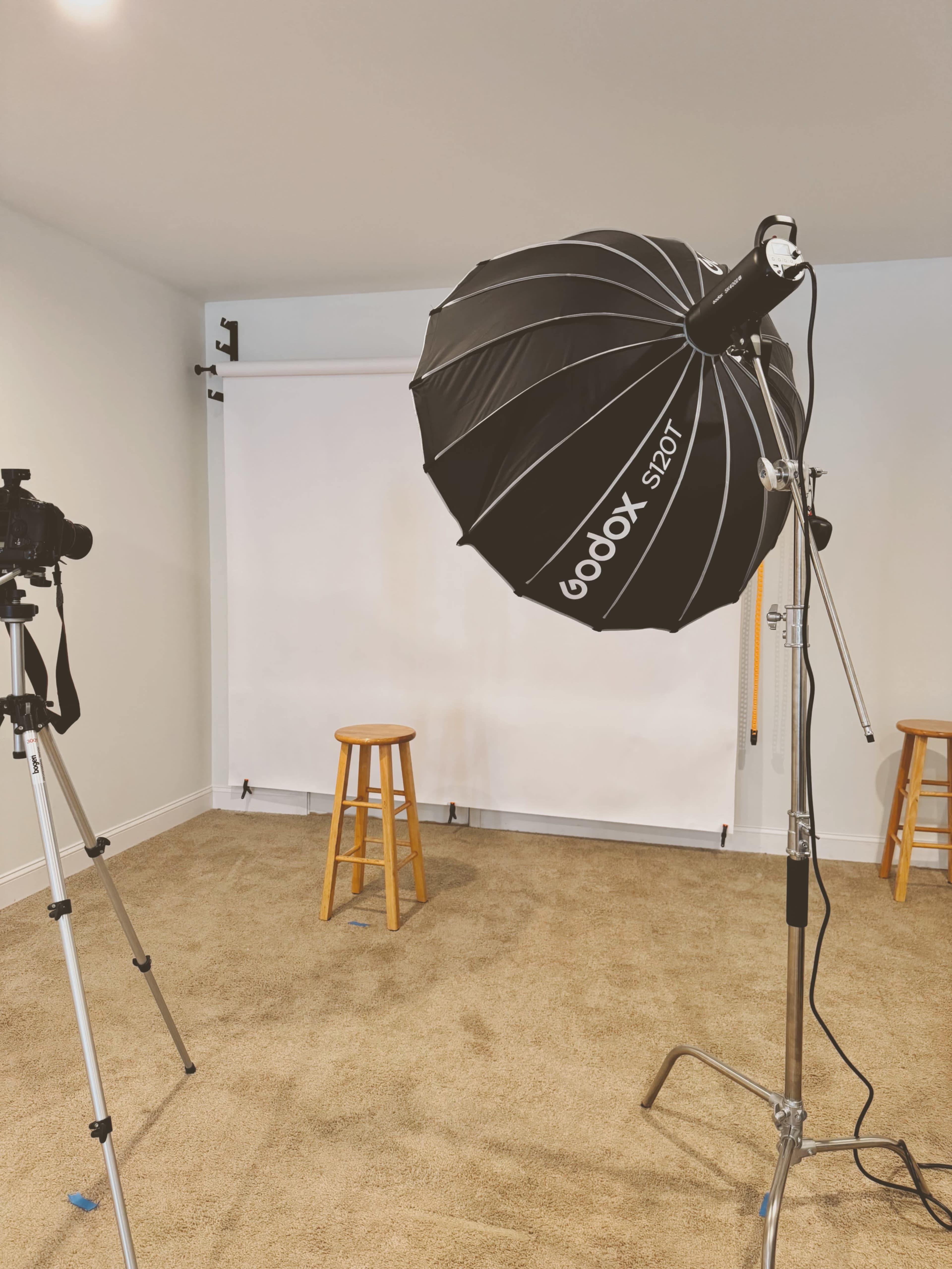 A photography setup is arranged in a room with a backdrop, a camera on a tripod, and an umbrella softbox light.