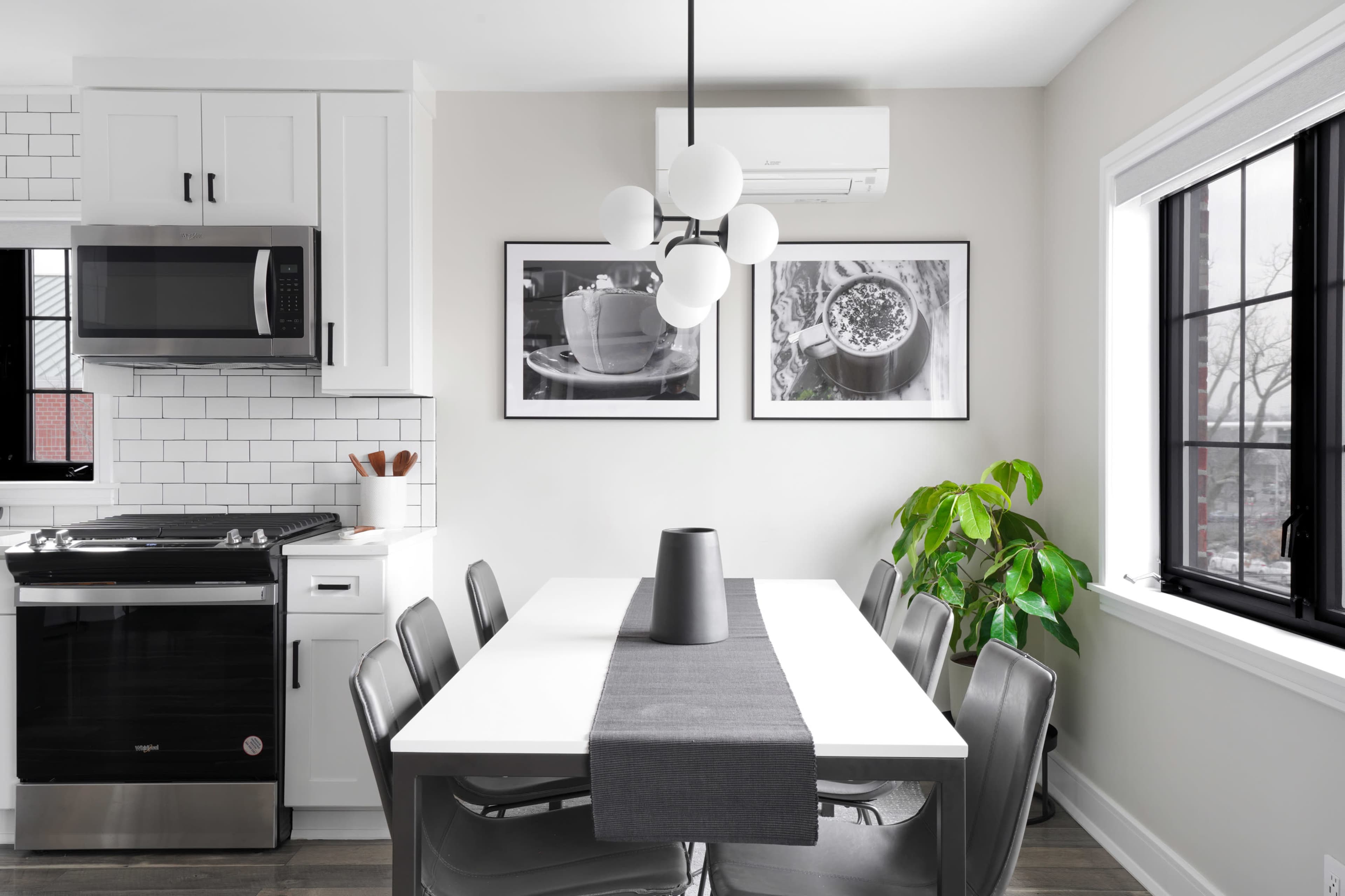 A modern kitchen features a dining table with black chairs, a large light fixture, and a potted plant near a window, complemented by black-and-white photos on the wall.