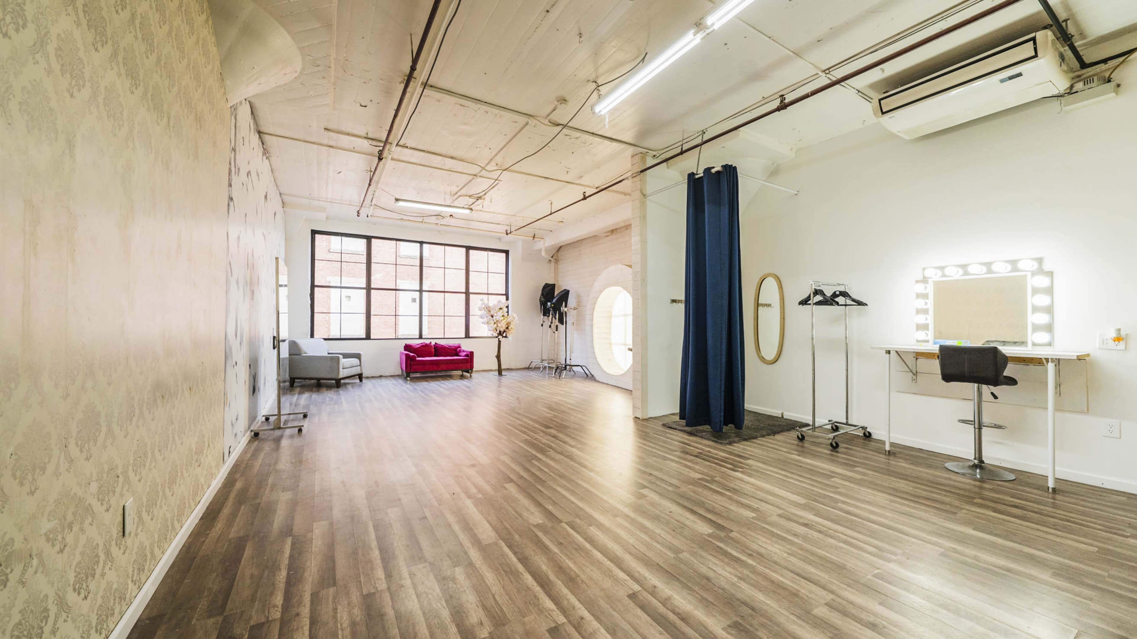 An open studio space with wooden flooring, a decorative wall, a large window, a seating area, and a makeup station with a lit mirror.