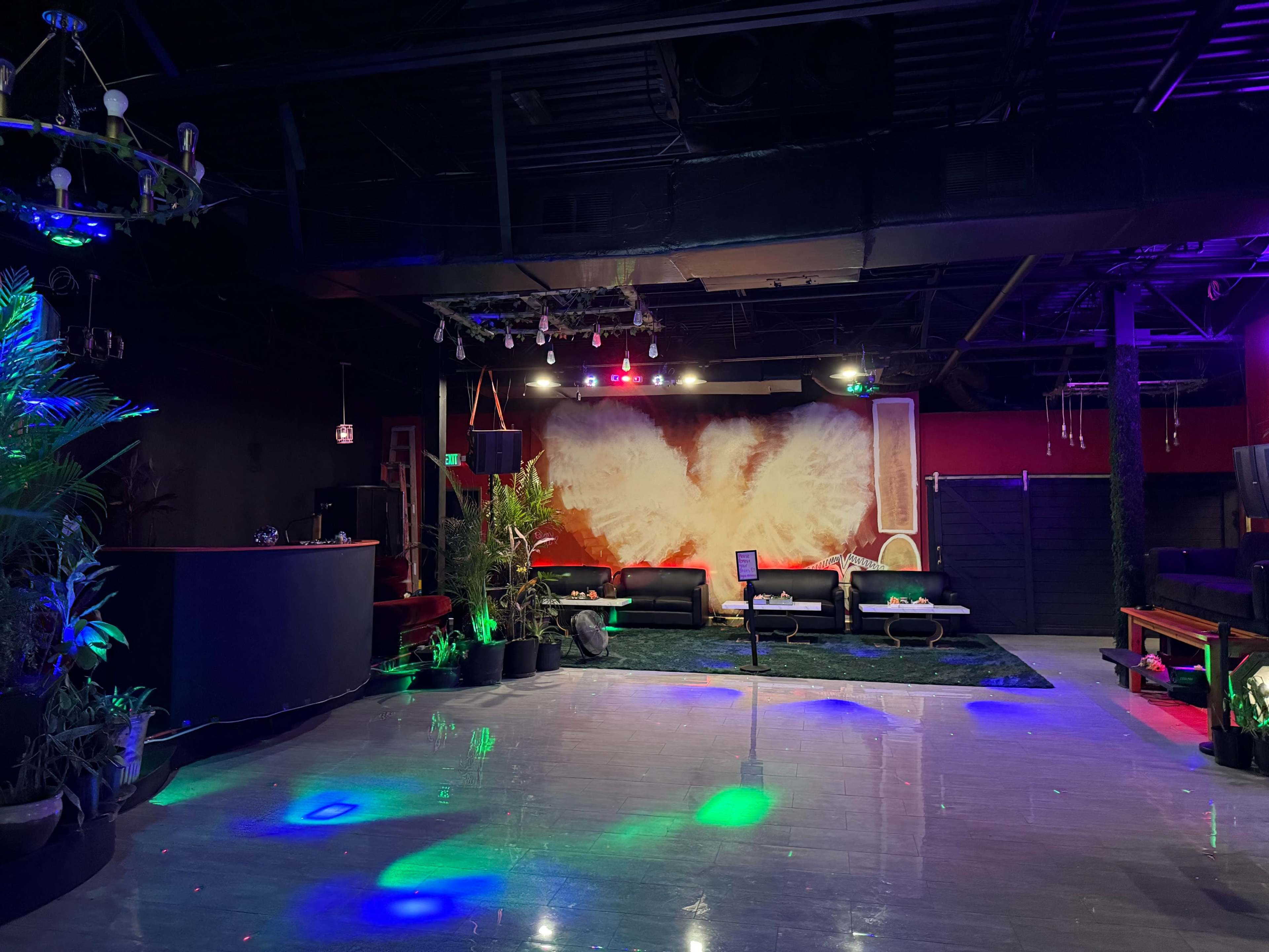 The image shows a spacious lounge area with a bar counter, several couches, and a large mural featuring wings on the back wall, illuminated by colorful lights.