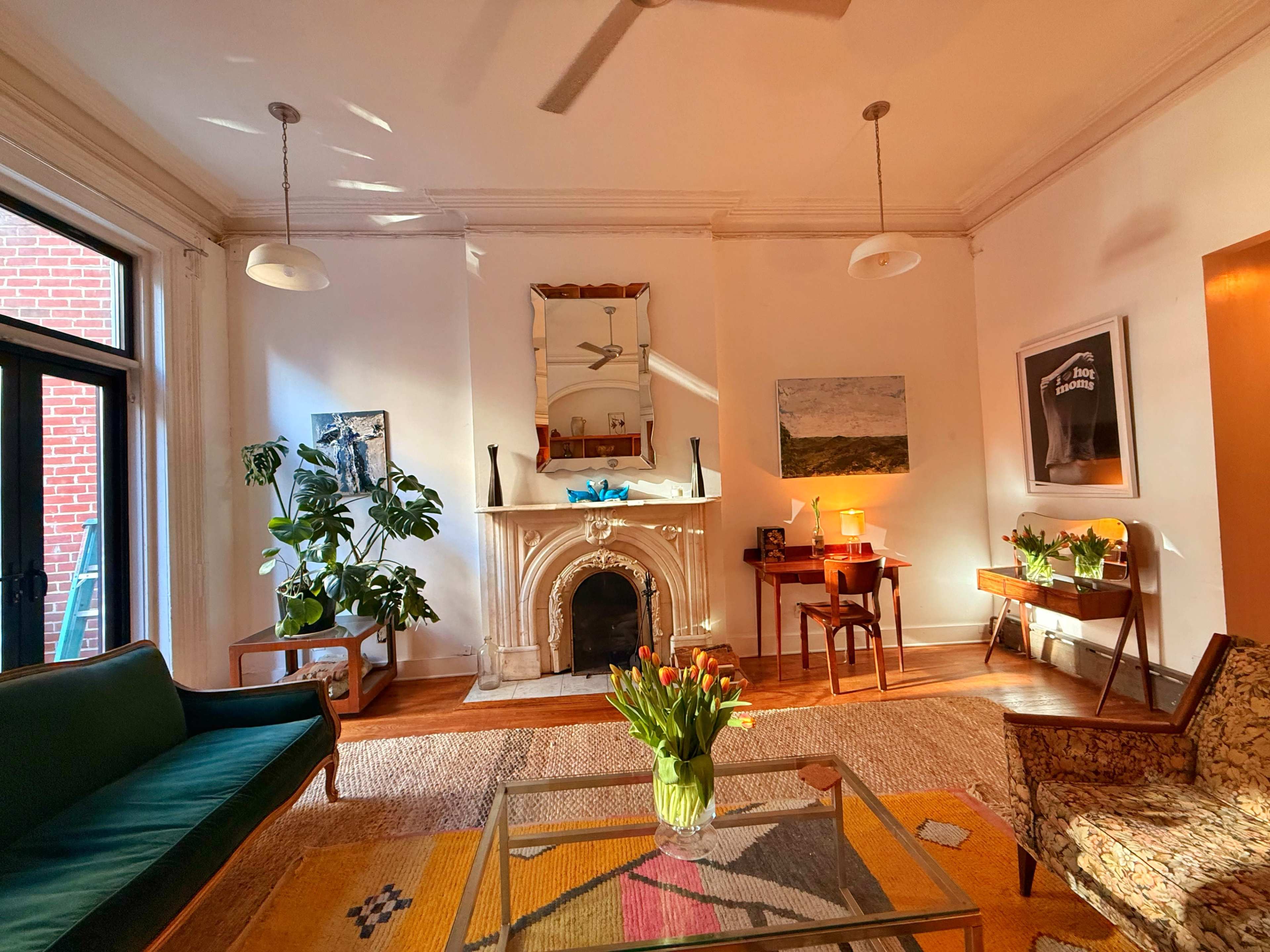 The image shows a well-lit living room featuring a green sofa, a patterned rug, an ornate fireplace, plants, and a small wooden desk with a lamp.
