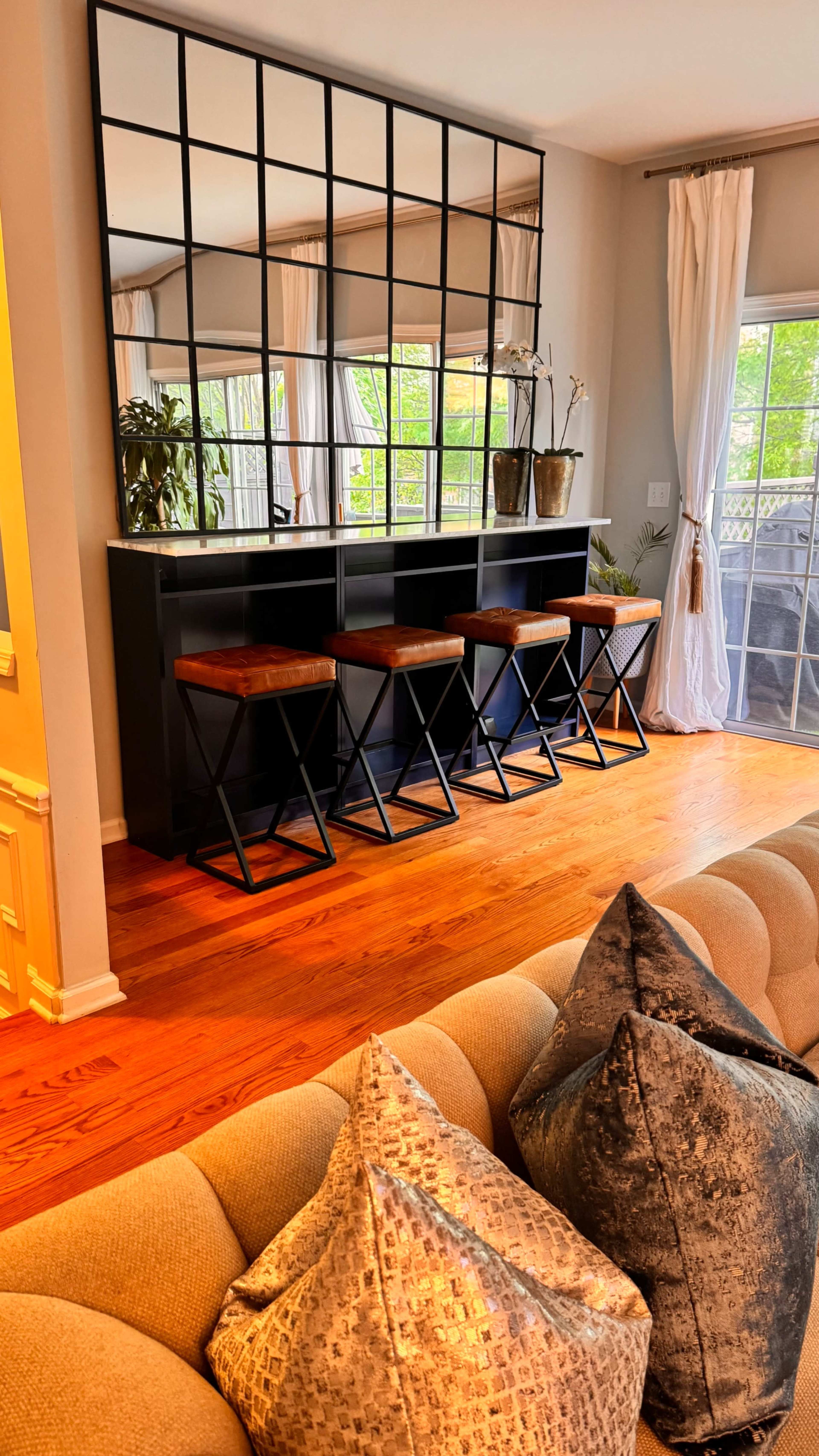 The image shows a living room with a sofa and a mirrored bar area featuring four high stools, decorated with potted plants and light-colored curtains.