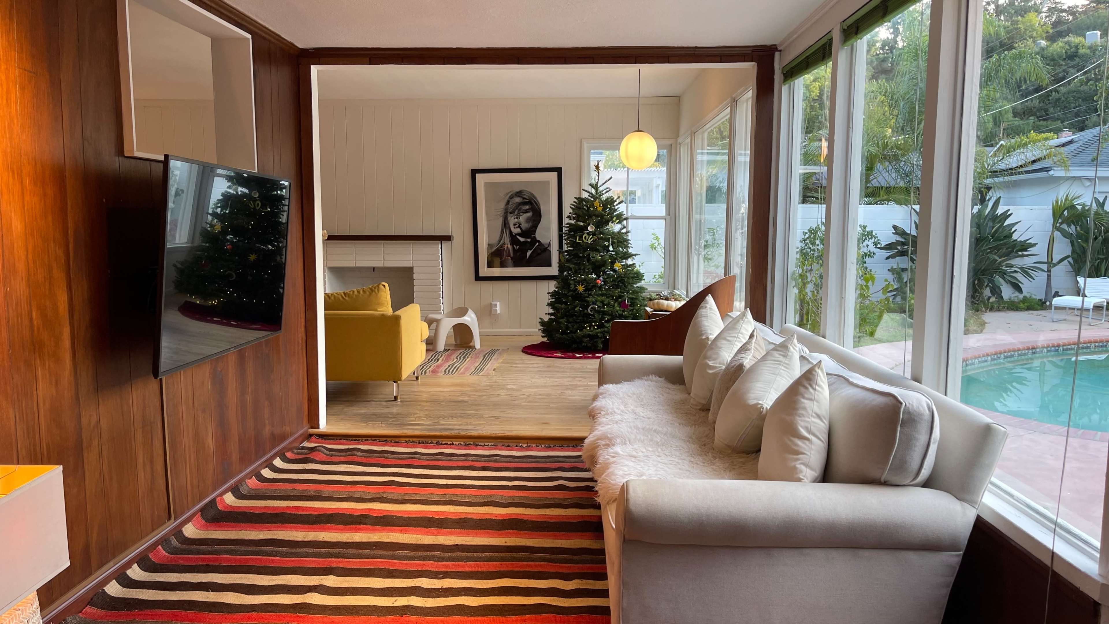 A cozy living room with a white sofa, a Christmas tree, large windows, and a yellow chair beside a swimming pool.