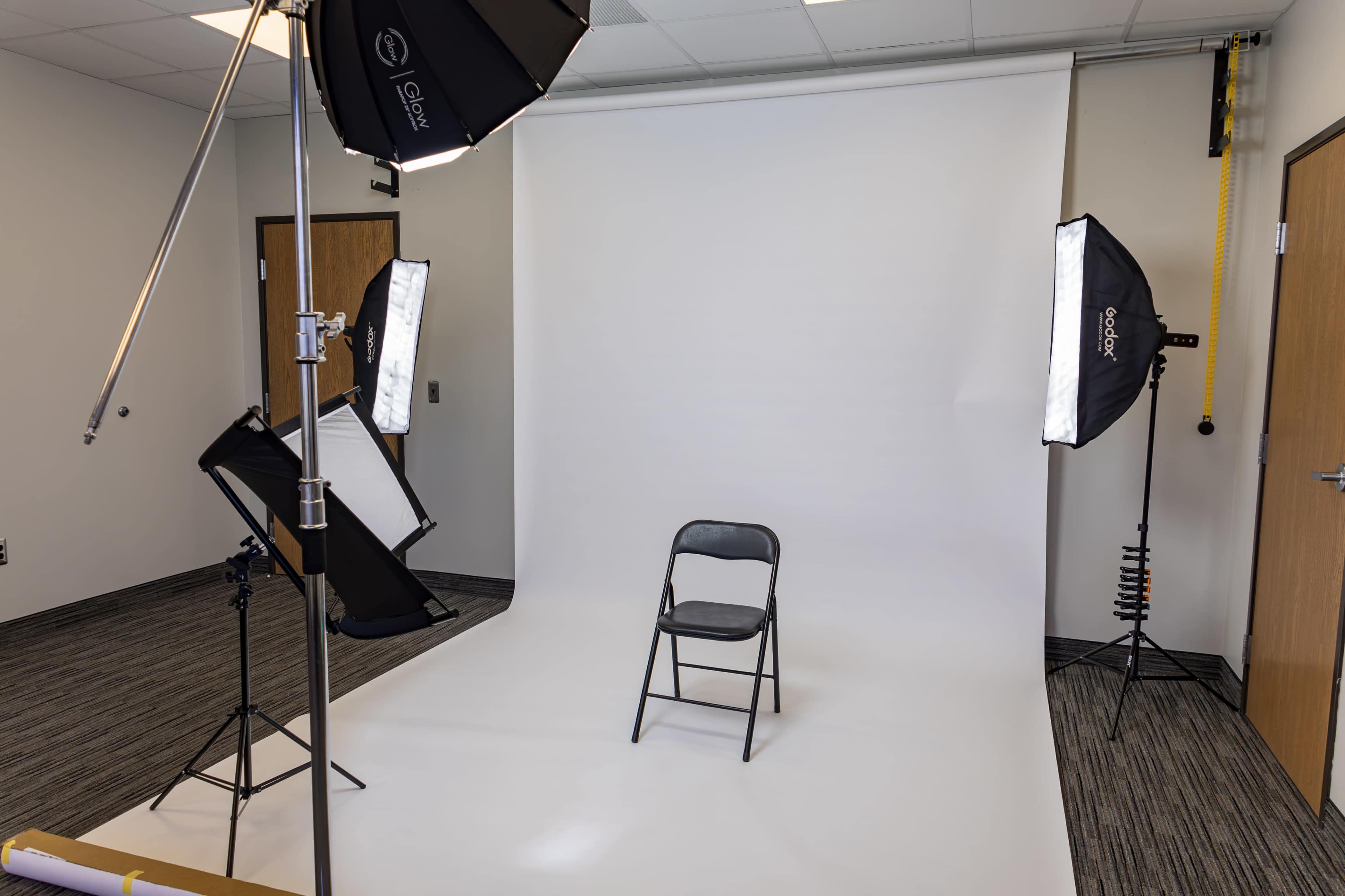 The image shows a photography studio setup with a single black chair positioned in front of a plain white backdrop, illuminated by softbox lights on either side.