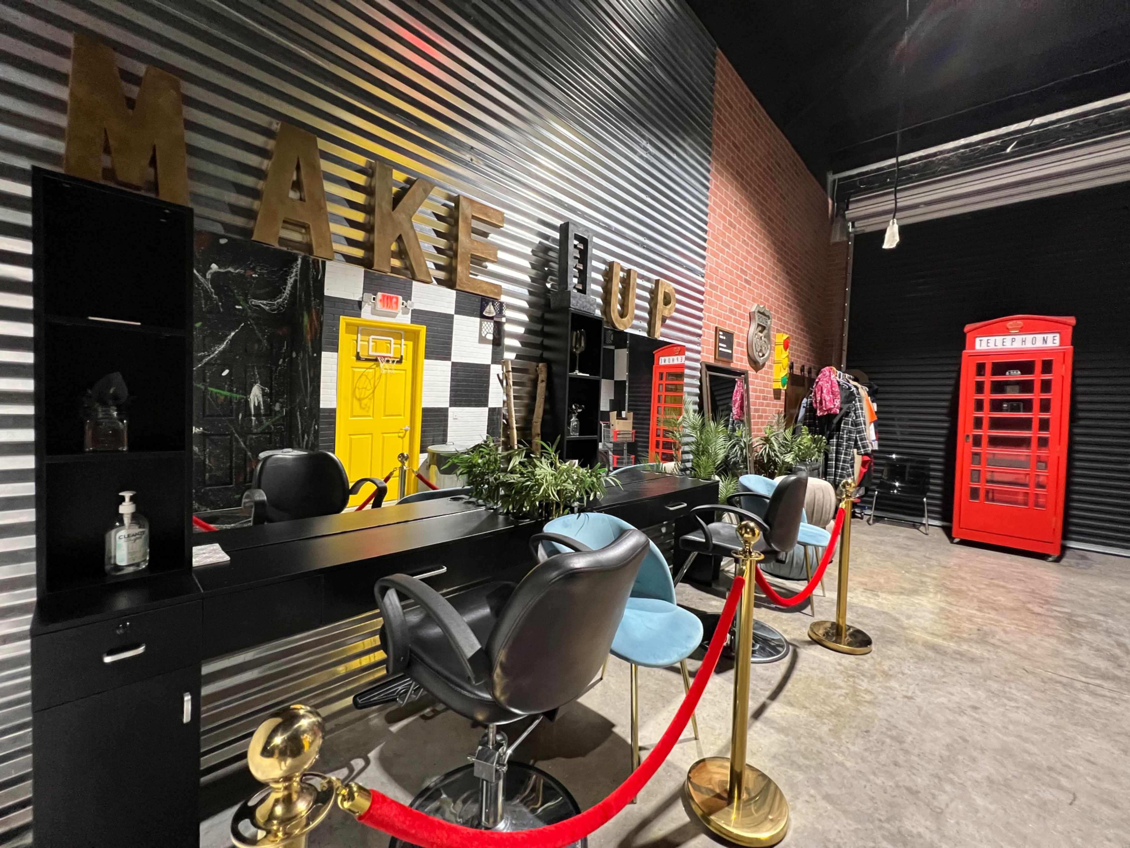 The image shows a makeup studio featuring a row of styling chairs, mirrors with large letters spelling "MAKEUP," potted plants, and a red telephone booth in the background.