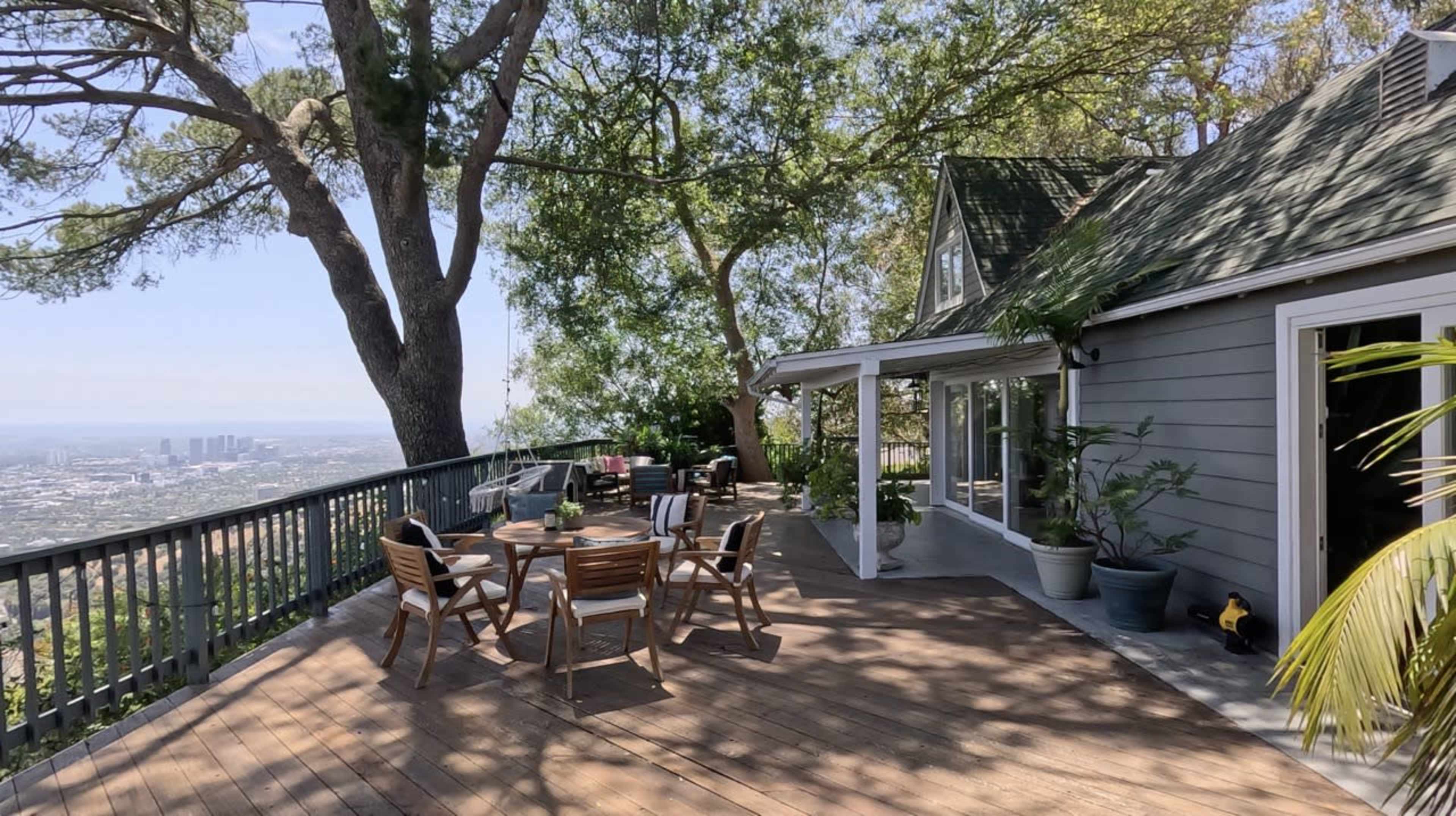 A spacious wooden deck with outdoor dining furniture overlooks a cityscape and is surrounded by greenery and a house with a sloped roof.