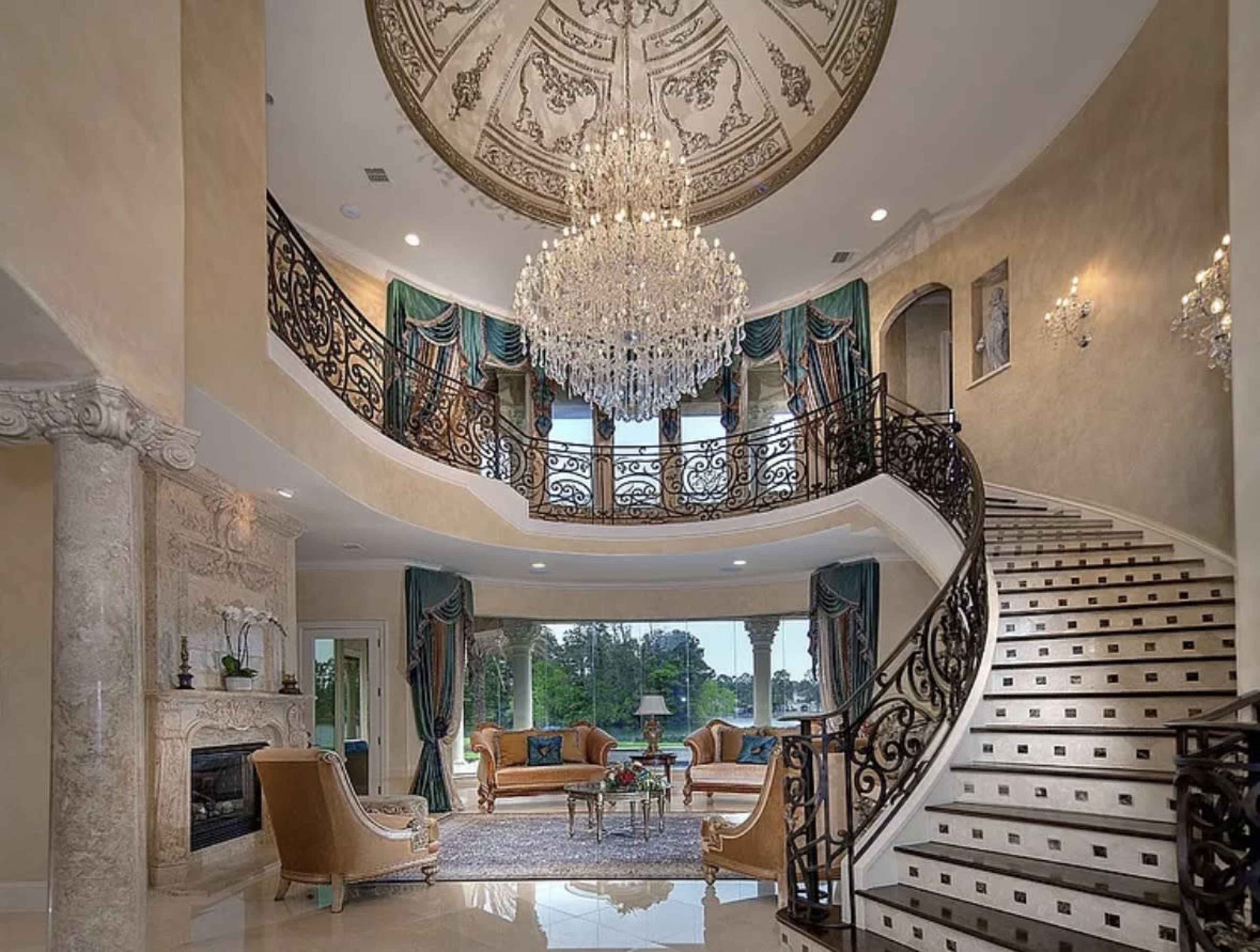 The image shows an elegantly designed foyer with a grand staircase, ornate chandelier, and decorative ceiling details, complemented by luxurious furnishings and large windows that let in natural light.