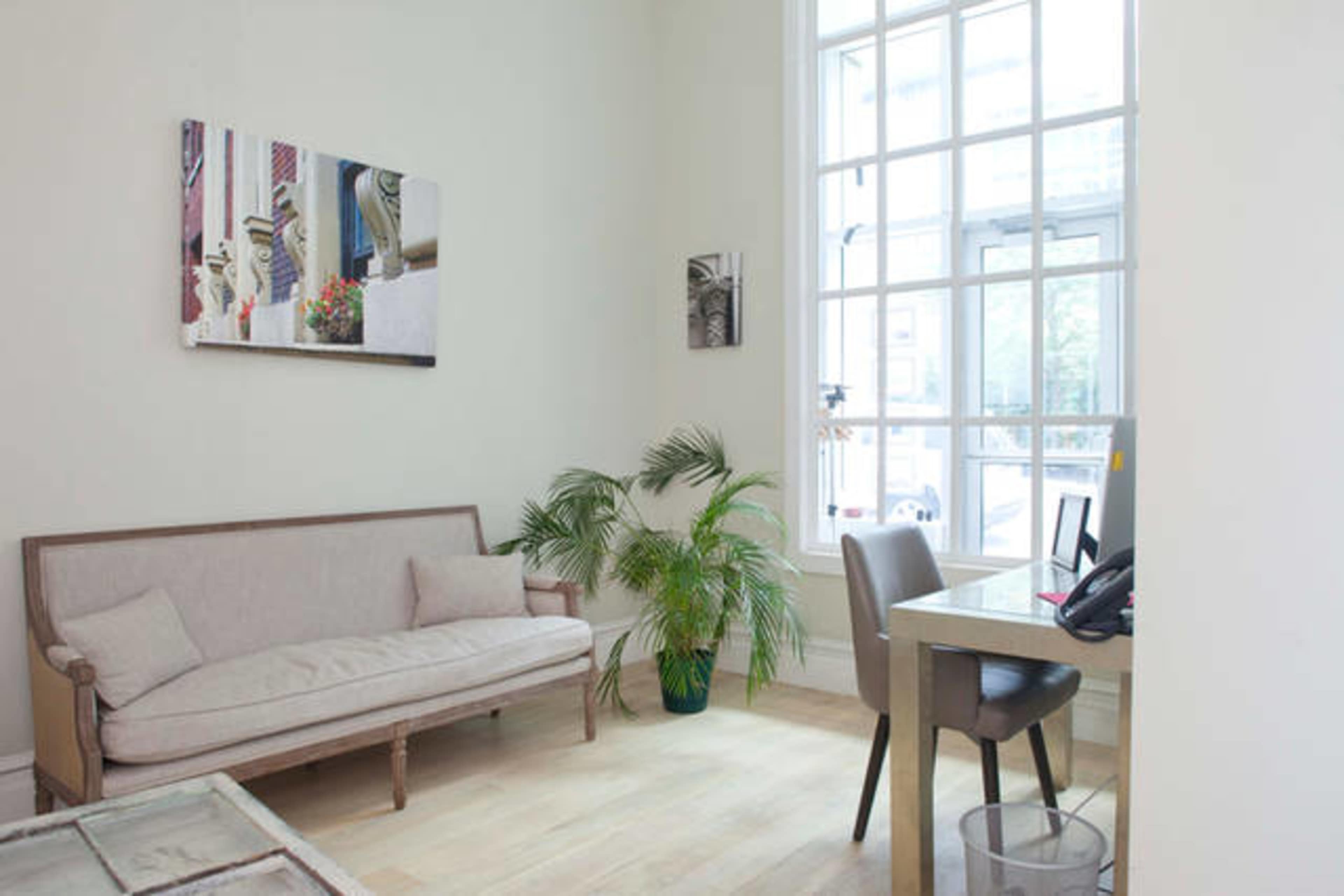 A light-filled room features a sofa, a desk with a chair, and decorative plants.
