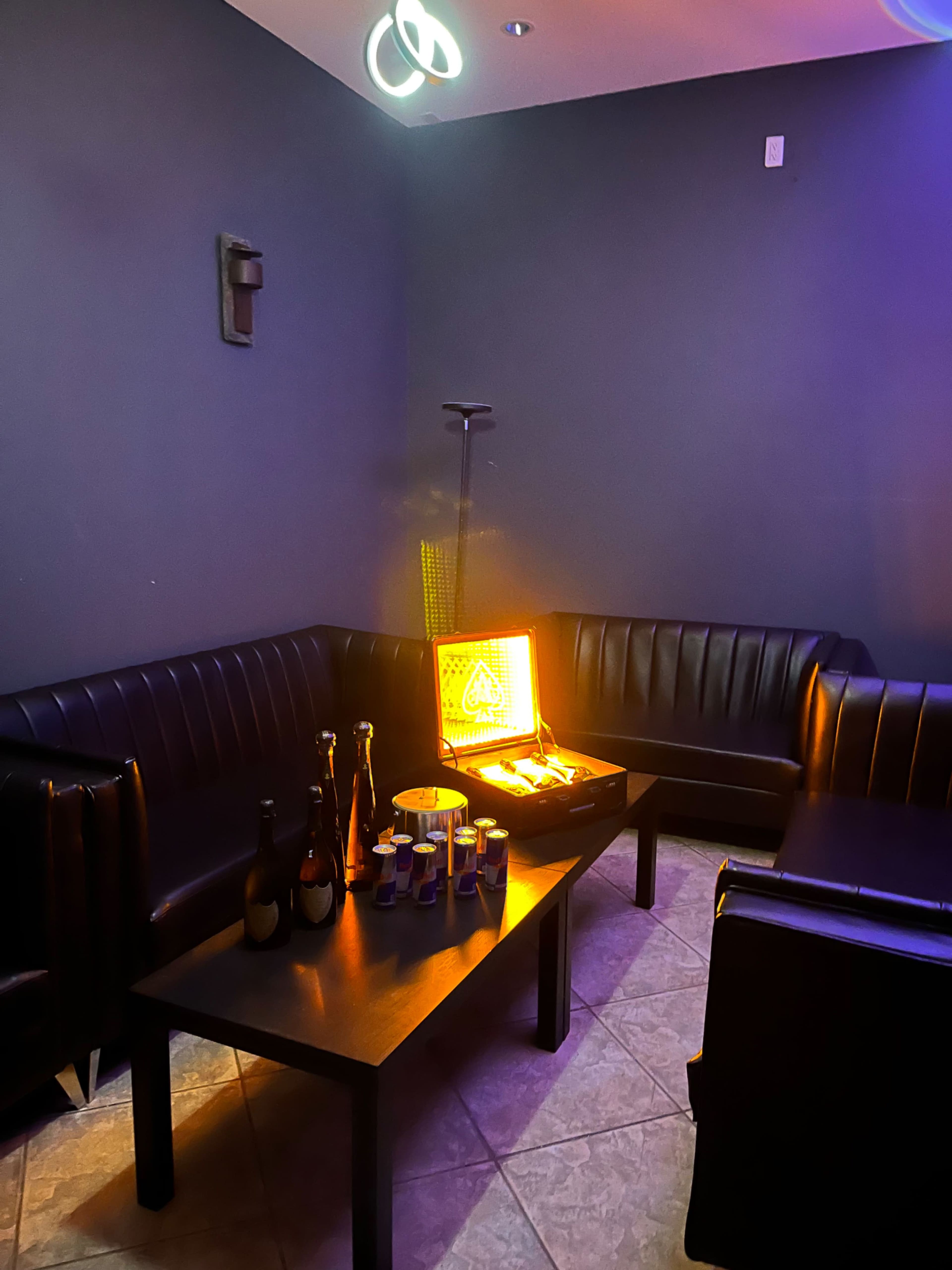 A dimly lit lounge area features black leather seating arranged around a low table with various bottles and a neon sign.