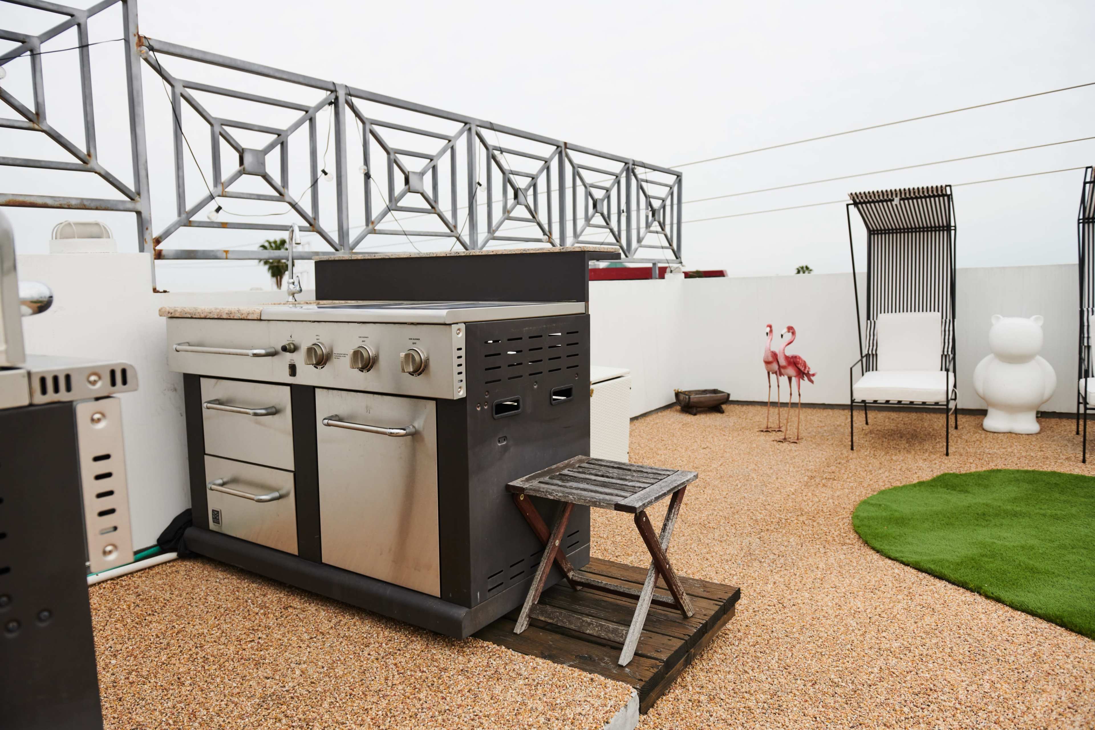 A rooftop terrace featuring a stainless steel barbecue grill, a small wooden table, a few chairs, and decorative elements like flamingos and a bear sculpture.