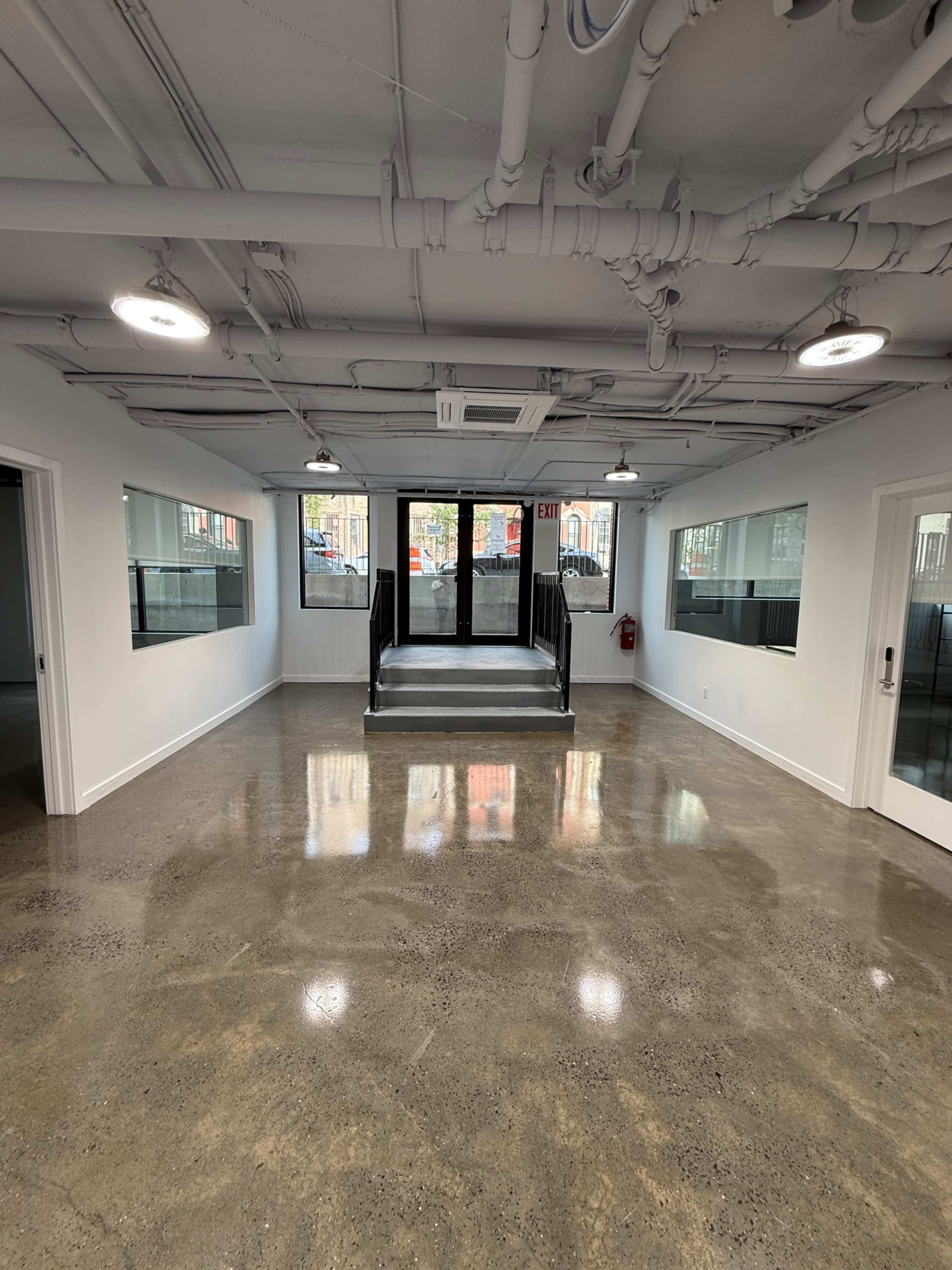 The image shows a spacious, industrial-style interior with polished concrete floors, exposed pipes on the ceiling, and large windows.