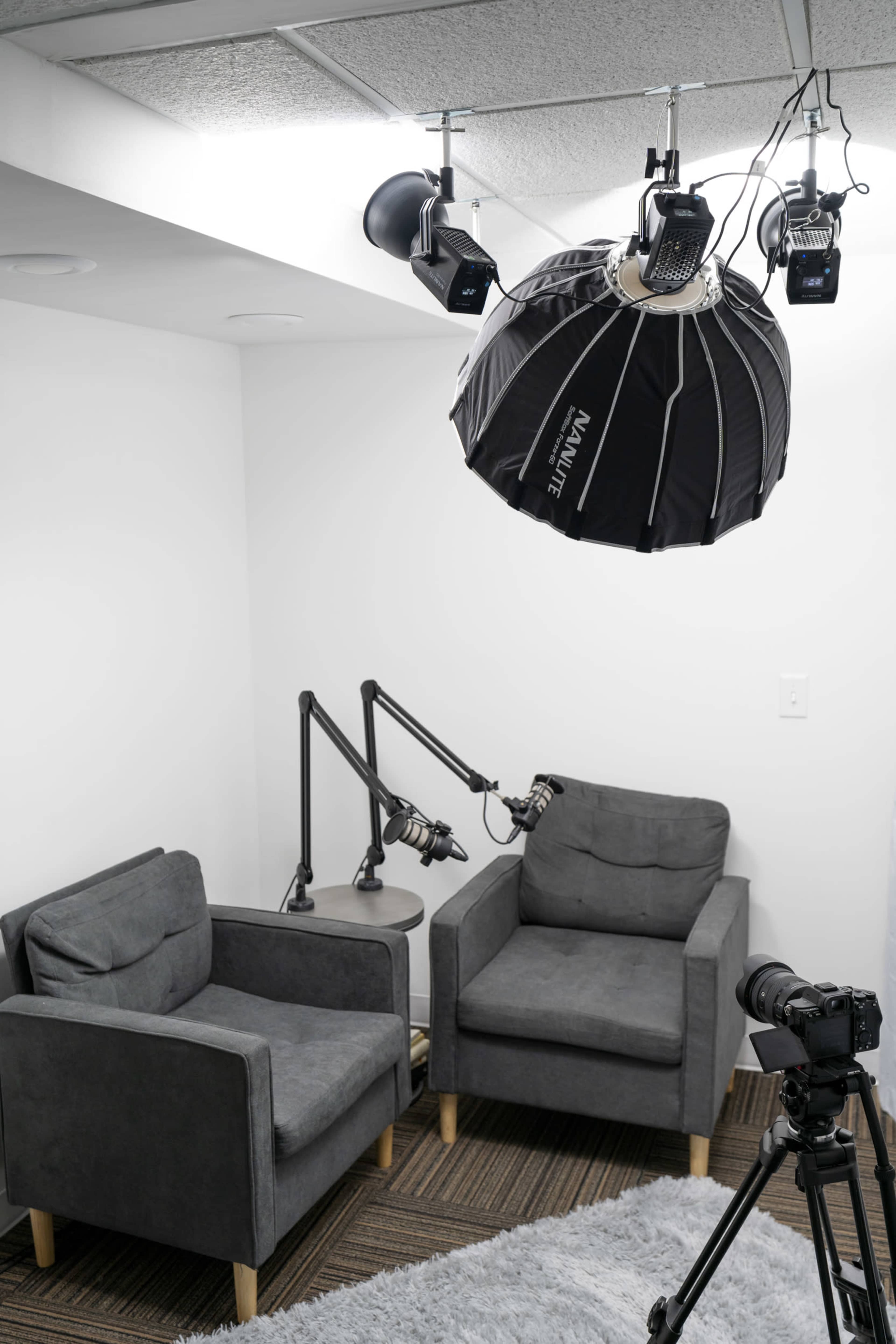A small recording studio featuring two gray chairs, a table, a large softbox light suspended from the ceiling, and a camera on a tripod.