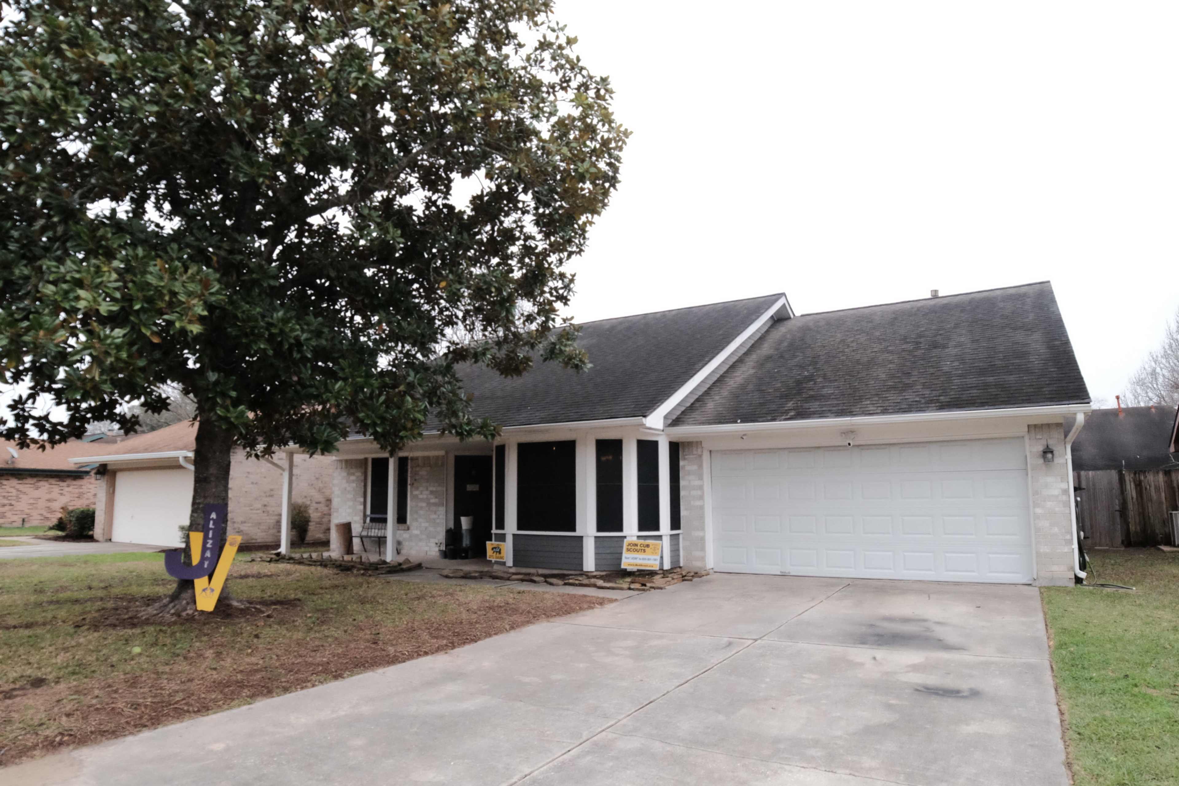 A single-story house with a sloped roof, a front porch, and a two-car garage is set on a grassy yard with a large tree on the left.
