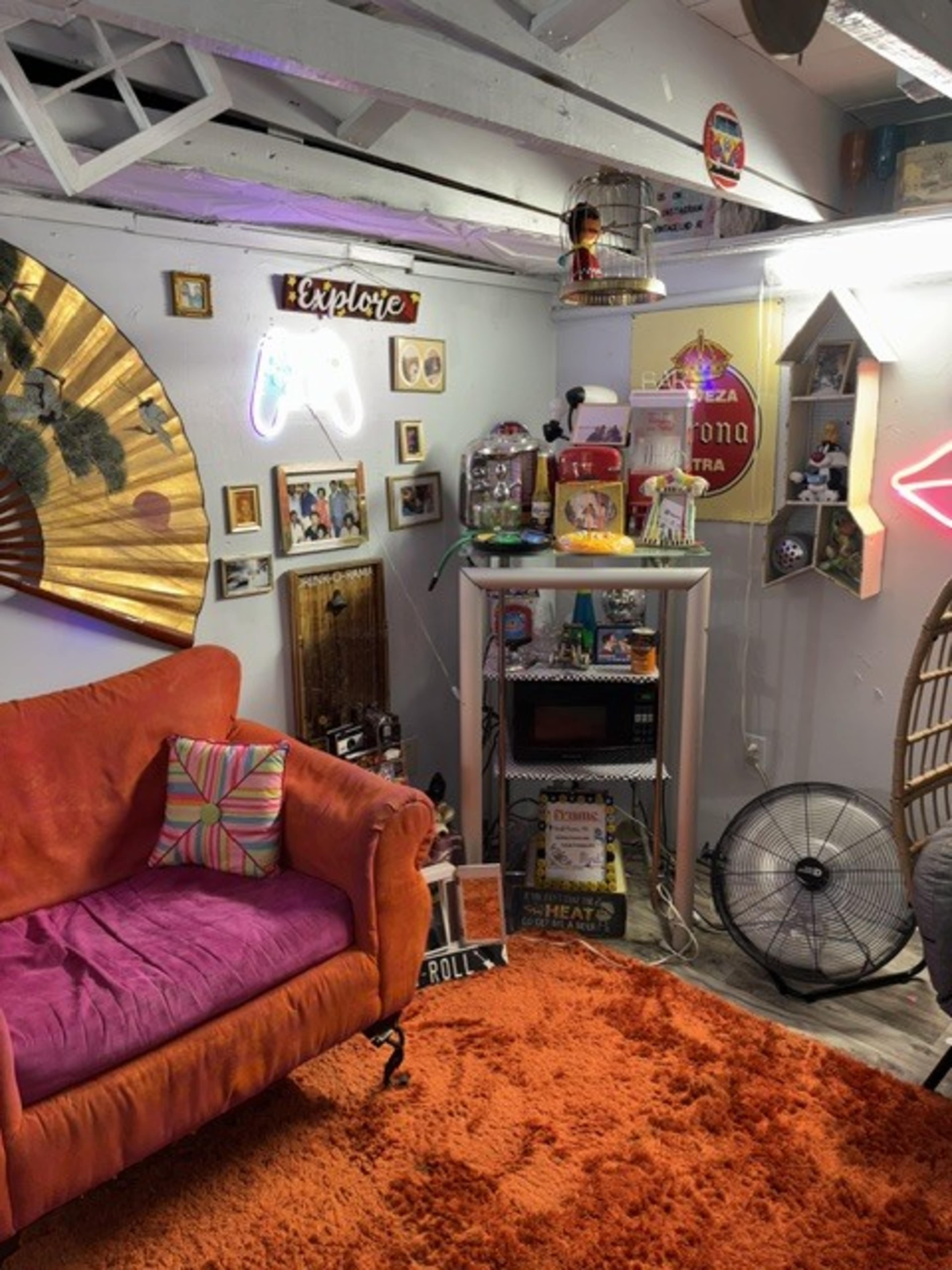 The image shows a colorful, eclectic room with a bright orange rug, a pink couch, various decorations on the walls, and a small shelf displaying knickknacks and a retro-style fan.