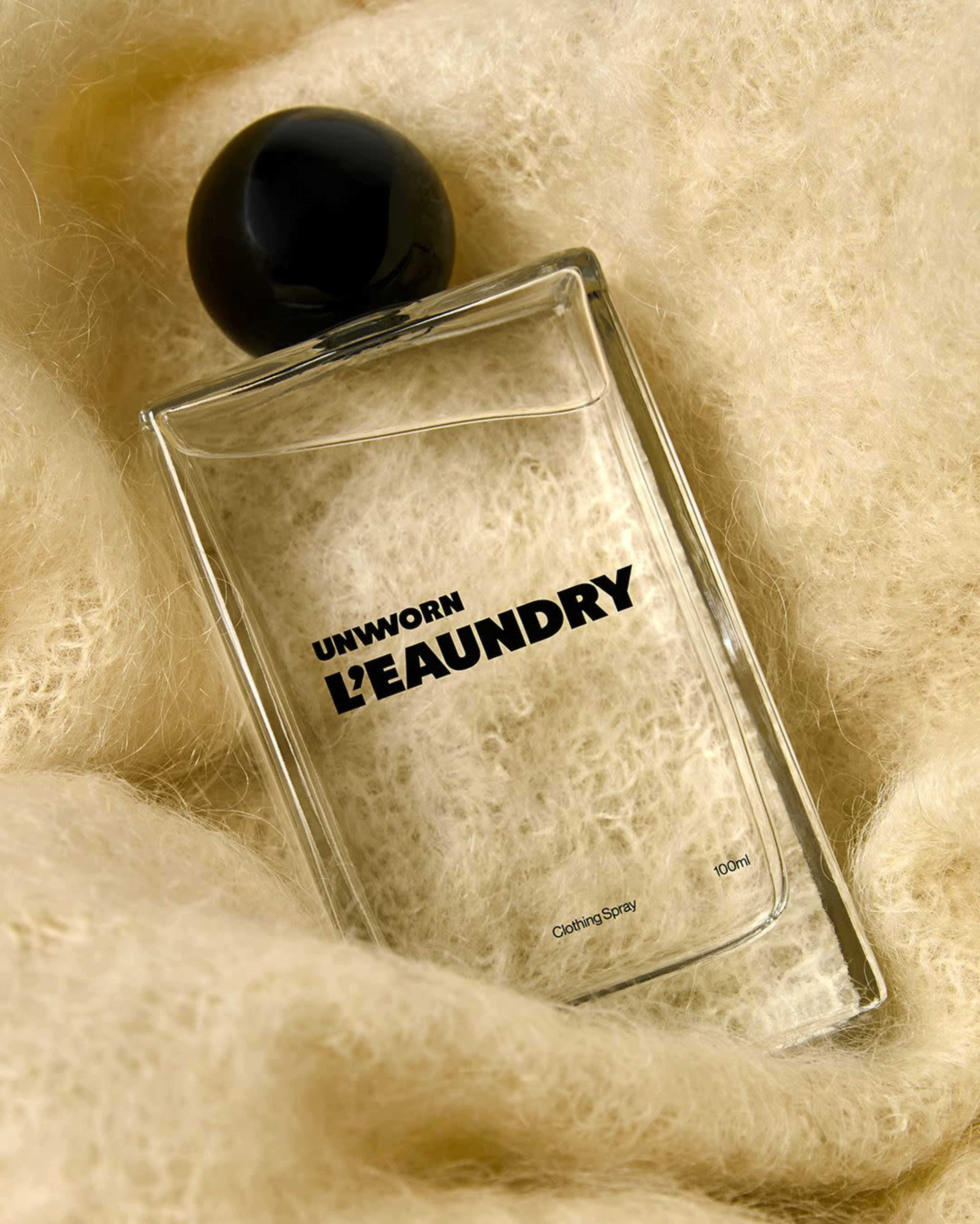 A clear perfume bottle with a black cap is placed on a soft, fluffy cream-colored background.