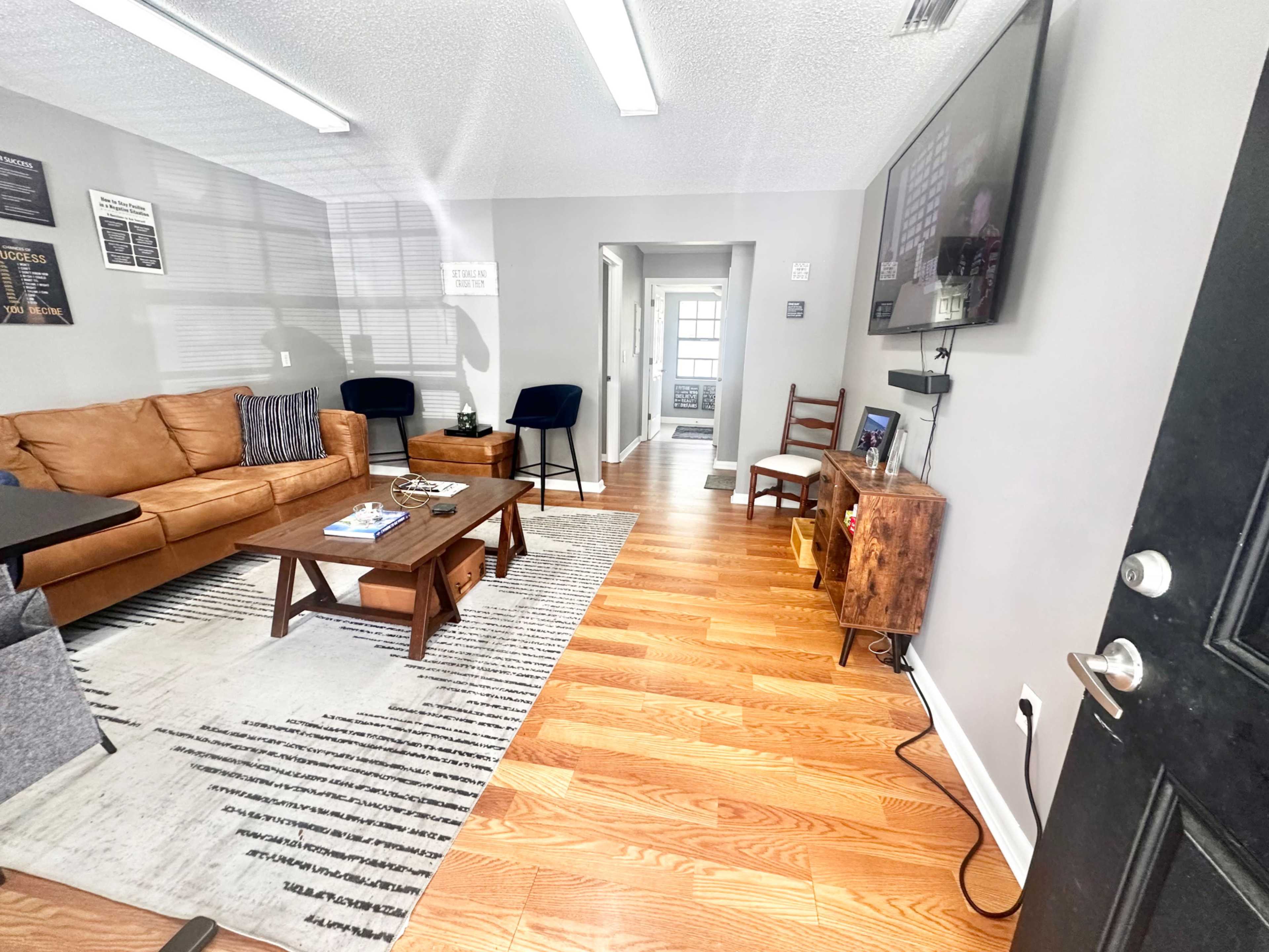 The image shows a modern living room with a brown leather sofa, a wooden coffee table, and a flat-screen TV mounted on the wall, featuring hardwood floors and neutral-colored walls.