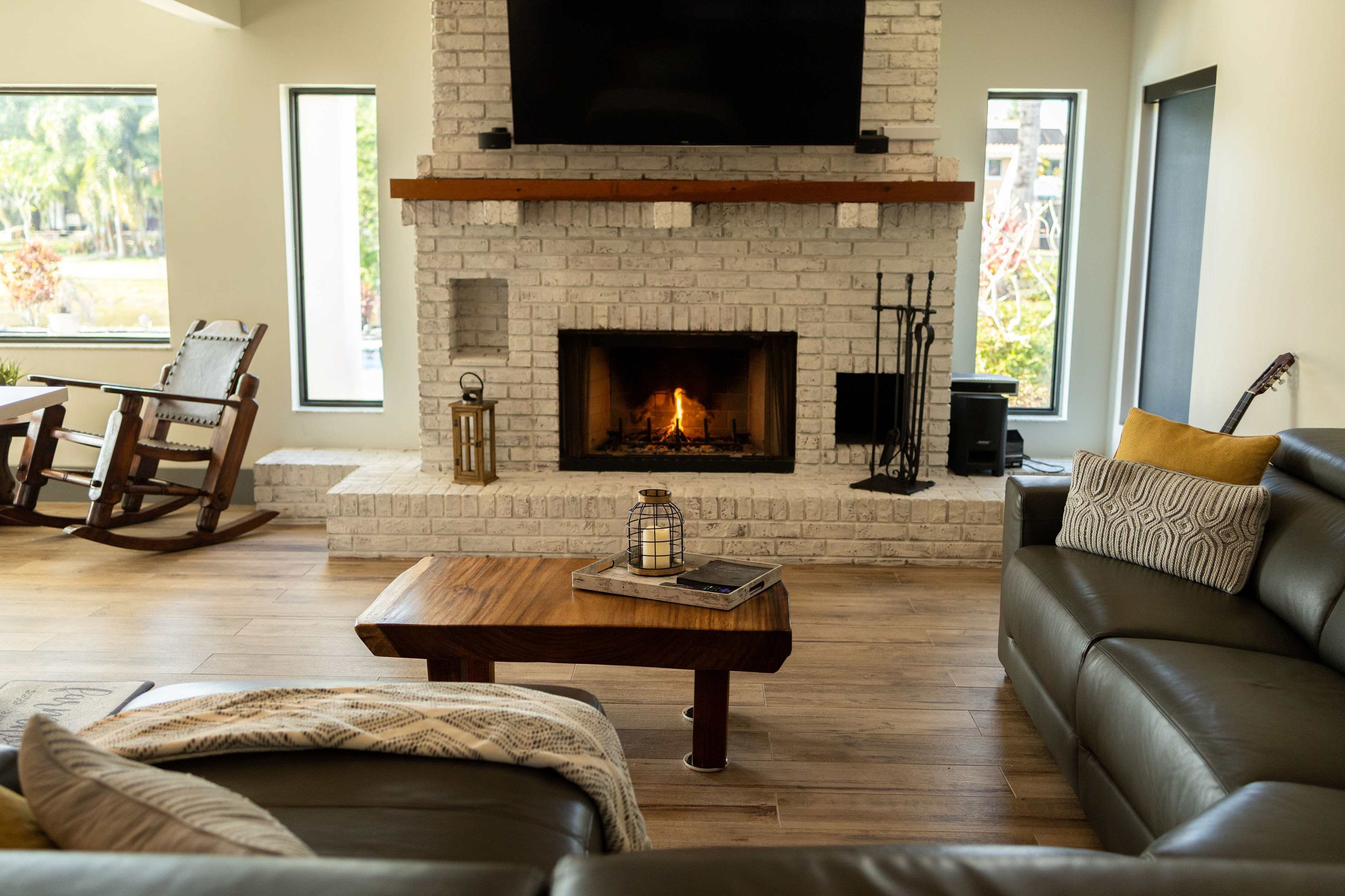 A modern living room features a stone fireplace with a fire burning, flanked by large windows and furnished with a leather sofa, wooden coffee table, and a rocking chair.