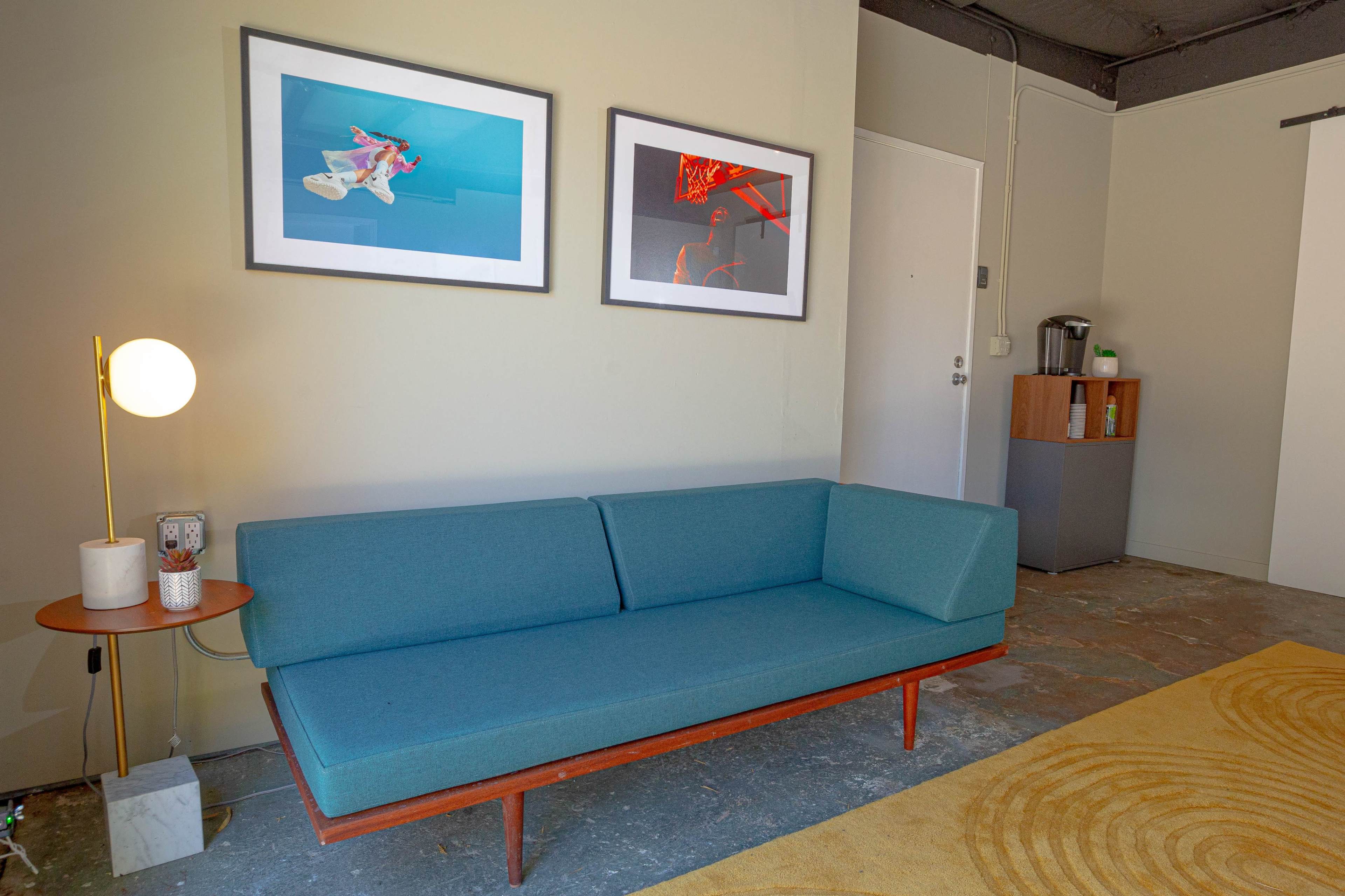 A modern living space featuring a blue upholstered sofa, two framed artworks on the wall, a round side table with a lamp, and a side cabinet with a coffee maker.