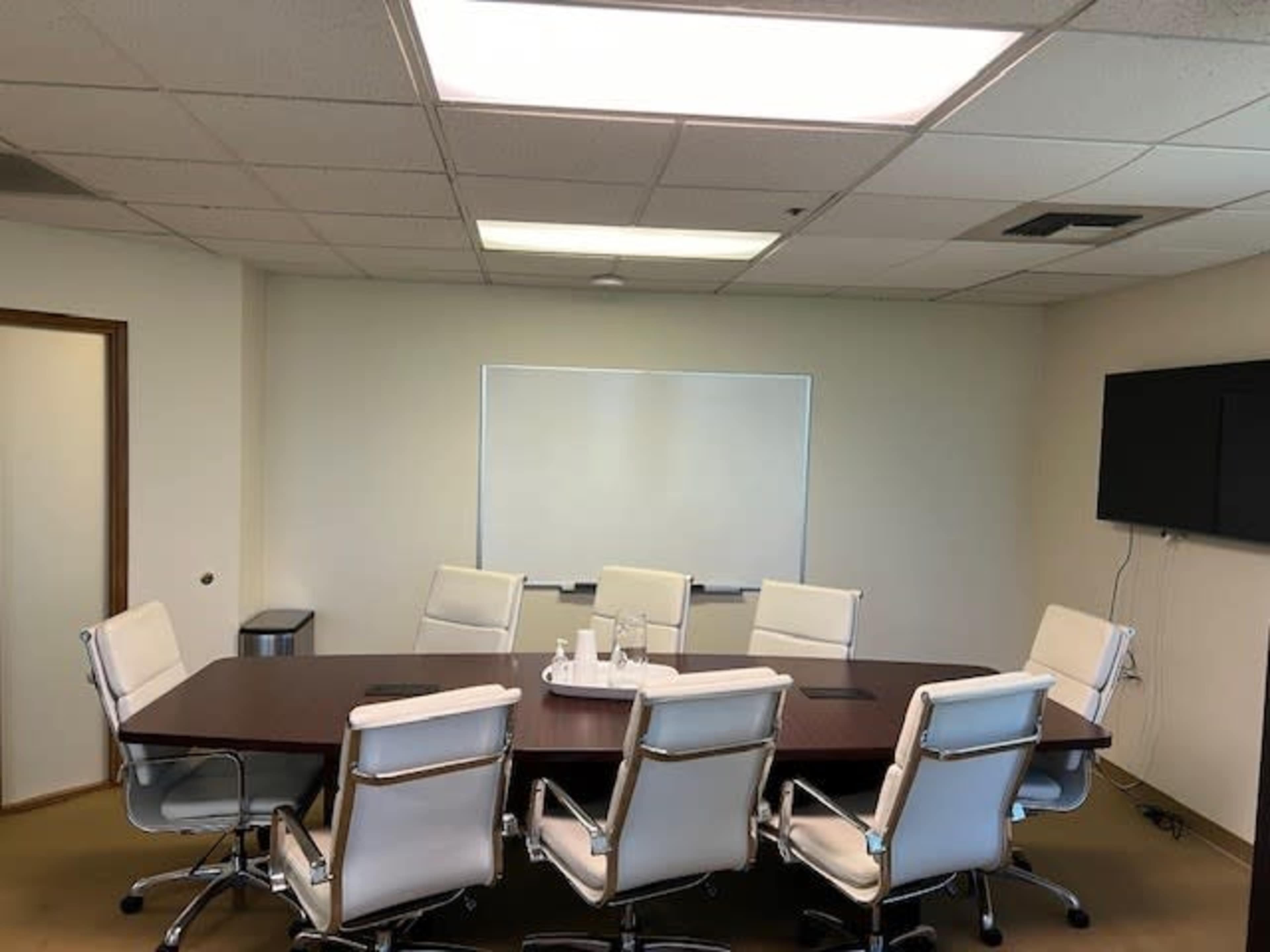 A conference room features a large rectangular table surrounded by eight white ergonomic chairs, a whiteboard on one wall, and a wall-mounted television on another.