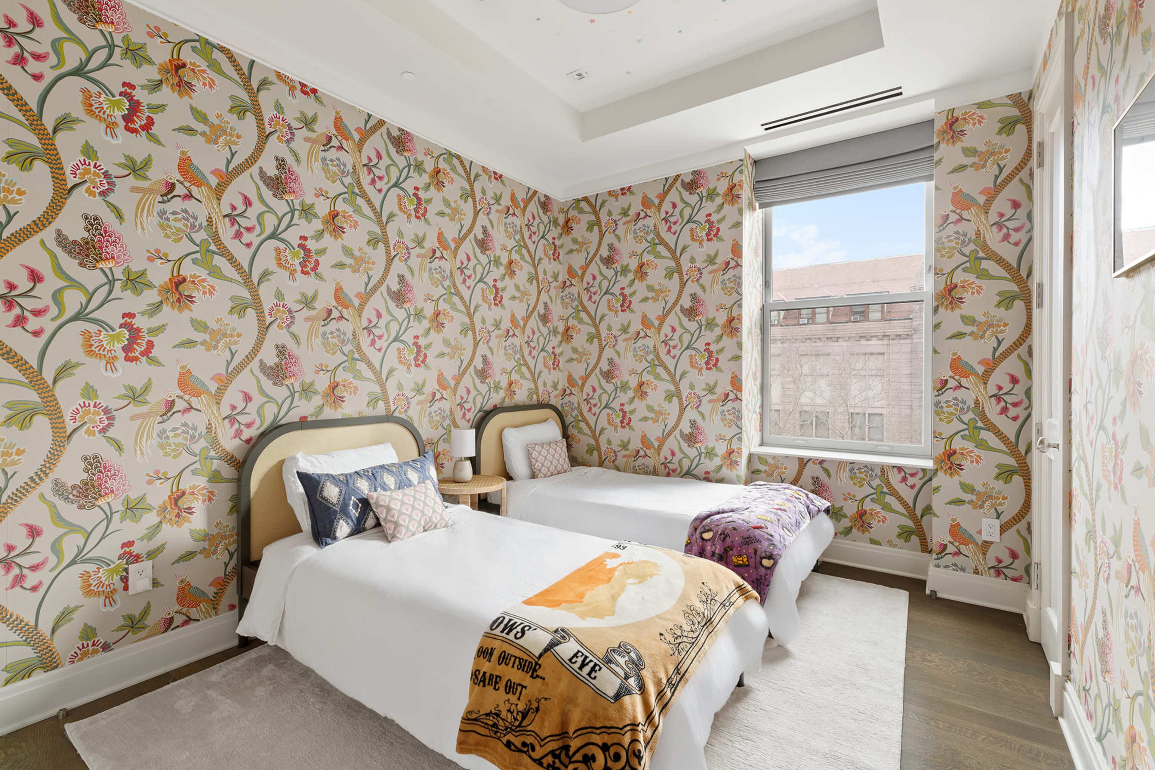 The image shows a brightly lit bedroom featuring two twin beds with decorative headboards and floral wallpaper.