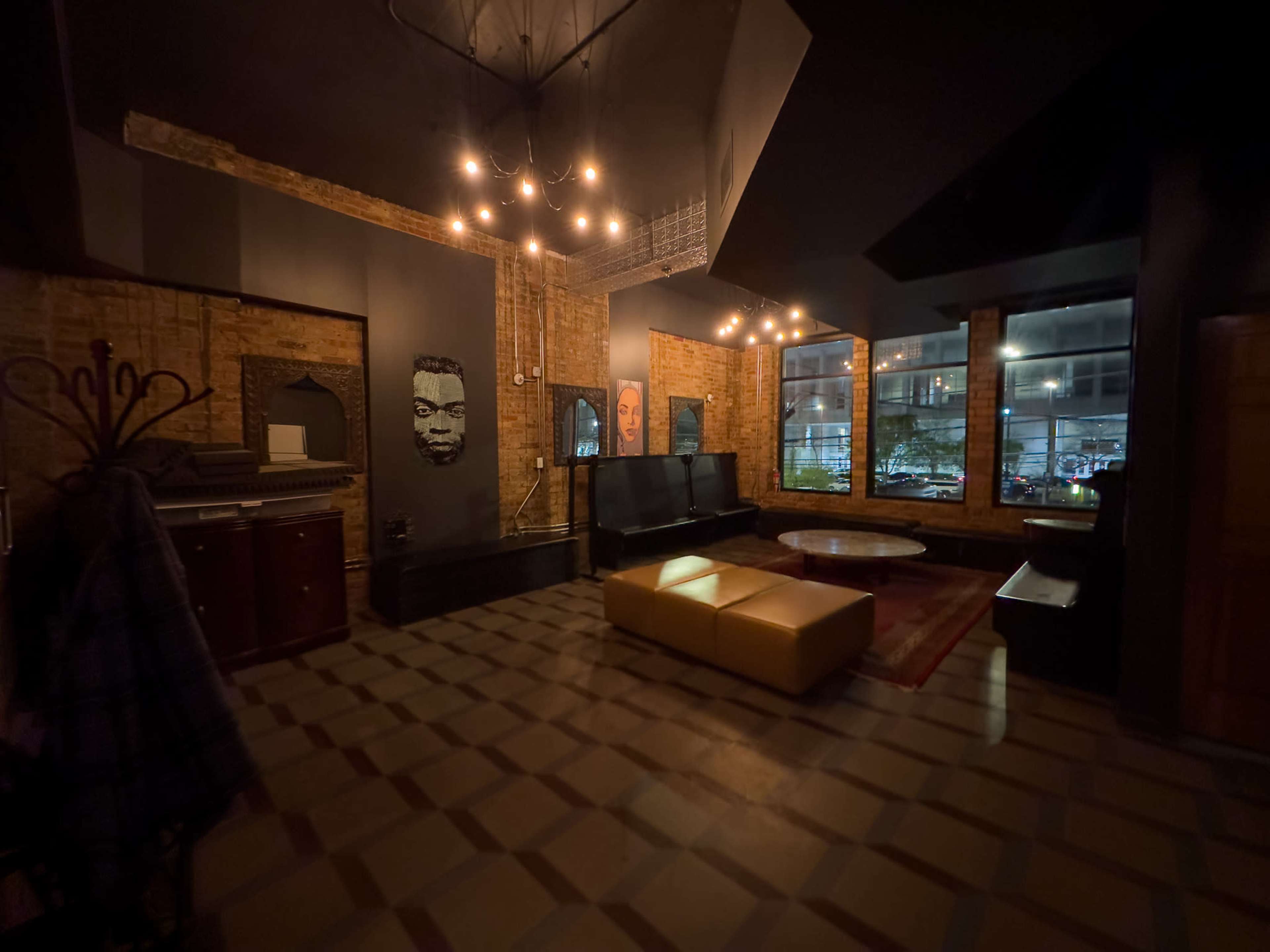 The image shows a dimly lit lounge featuring brick walls, modern lighting fixtures, and a mix of seating arrangements including a yellow sofa and dark chairs, with large windows revealing a nighttime city view.