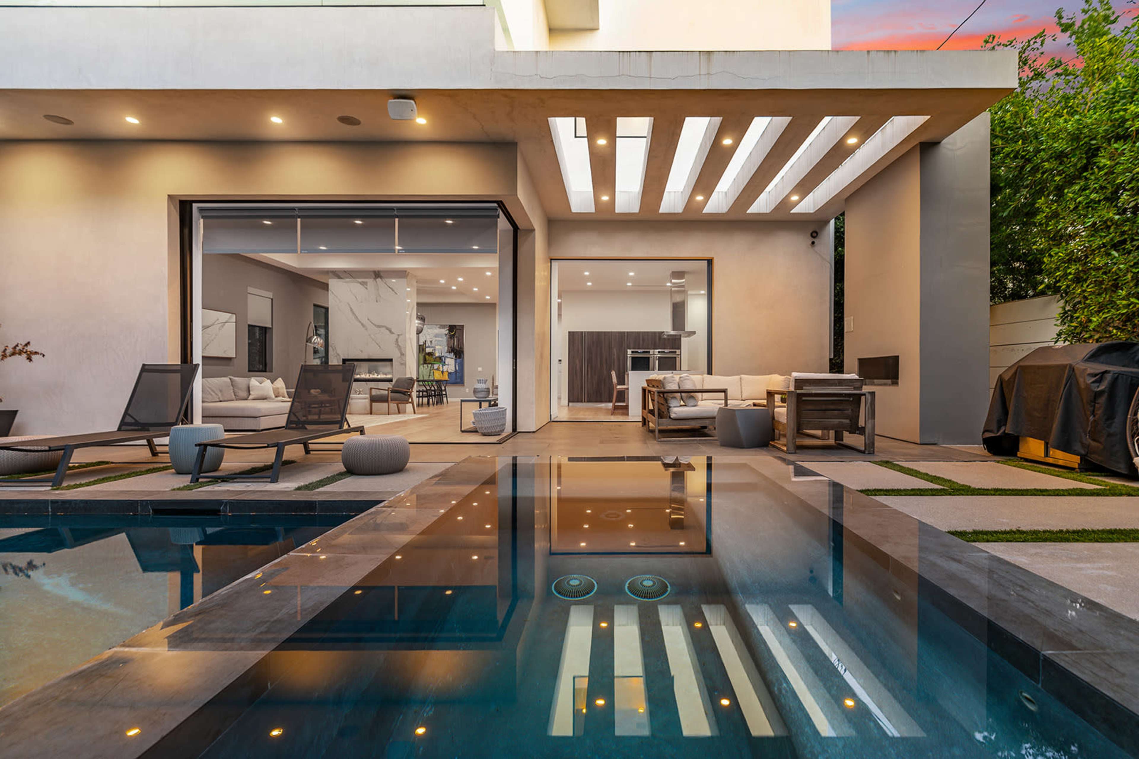 A modern outdoor space features a sleek pool reflecting the contemporary architecture of a home, with lounging chairs and a patio area visible.