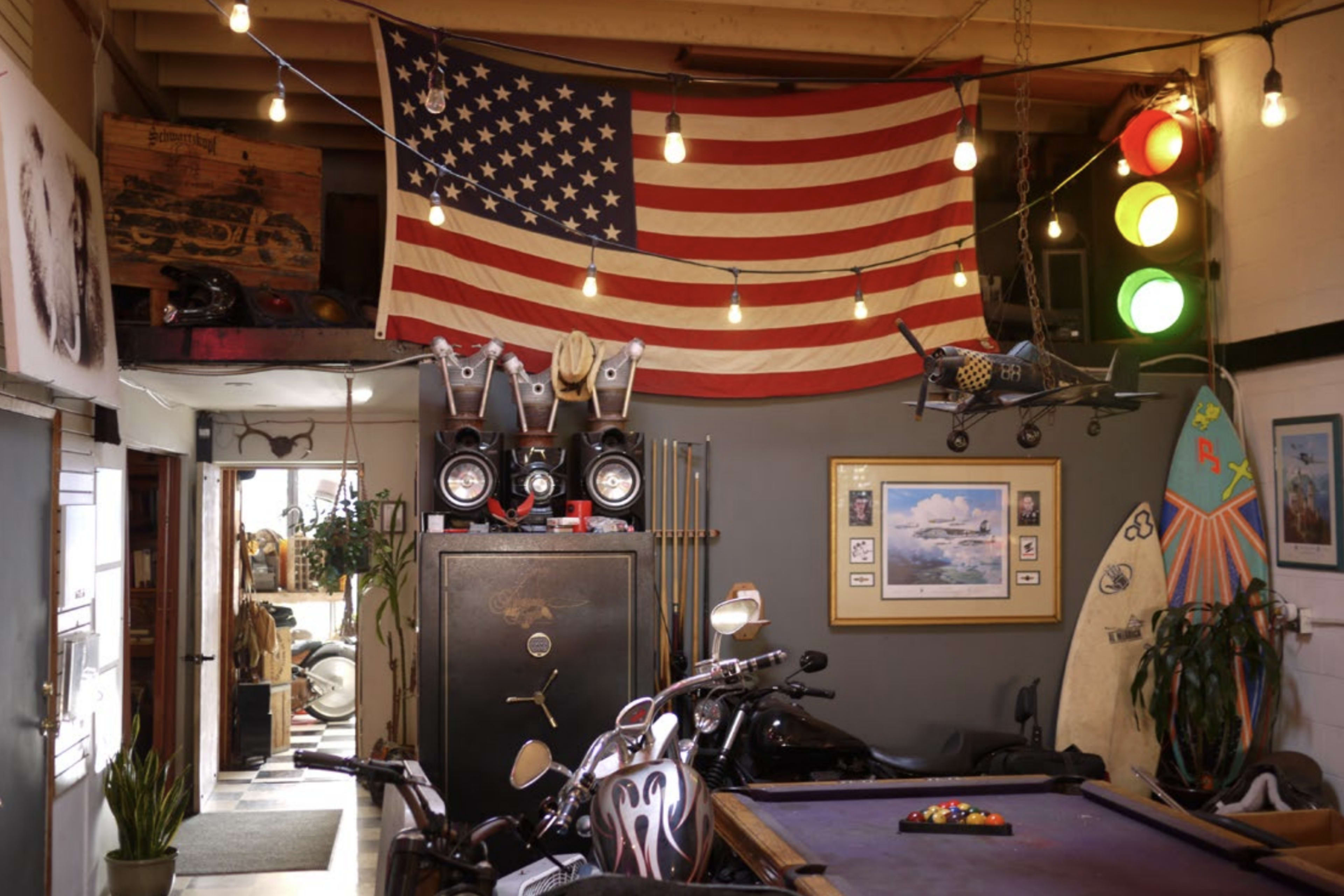 The image shows a garage or workshop featuring an American flag, a vintage safe, motorcycle parts, a pool table, and a surfboard, all illuminated by string lights.