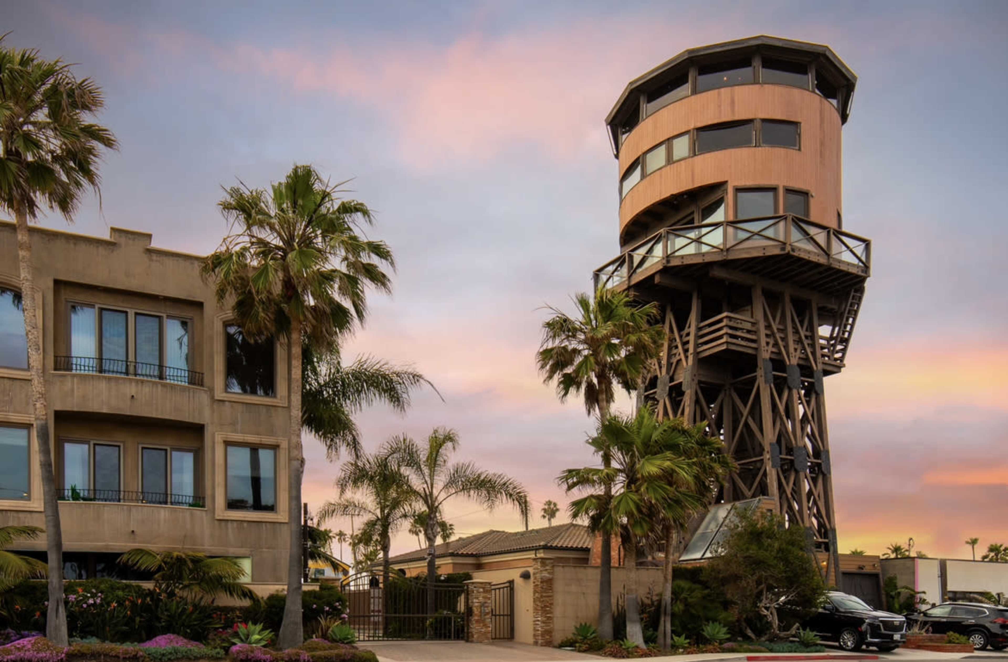 A tall observation tower made of wood and metal, surrounded by palm trees and a modern building, set against a colorful sunset sky.