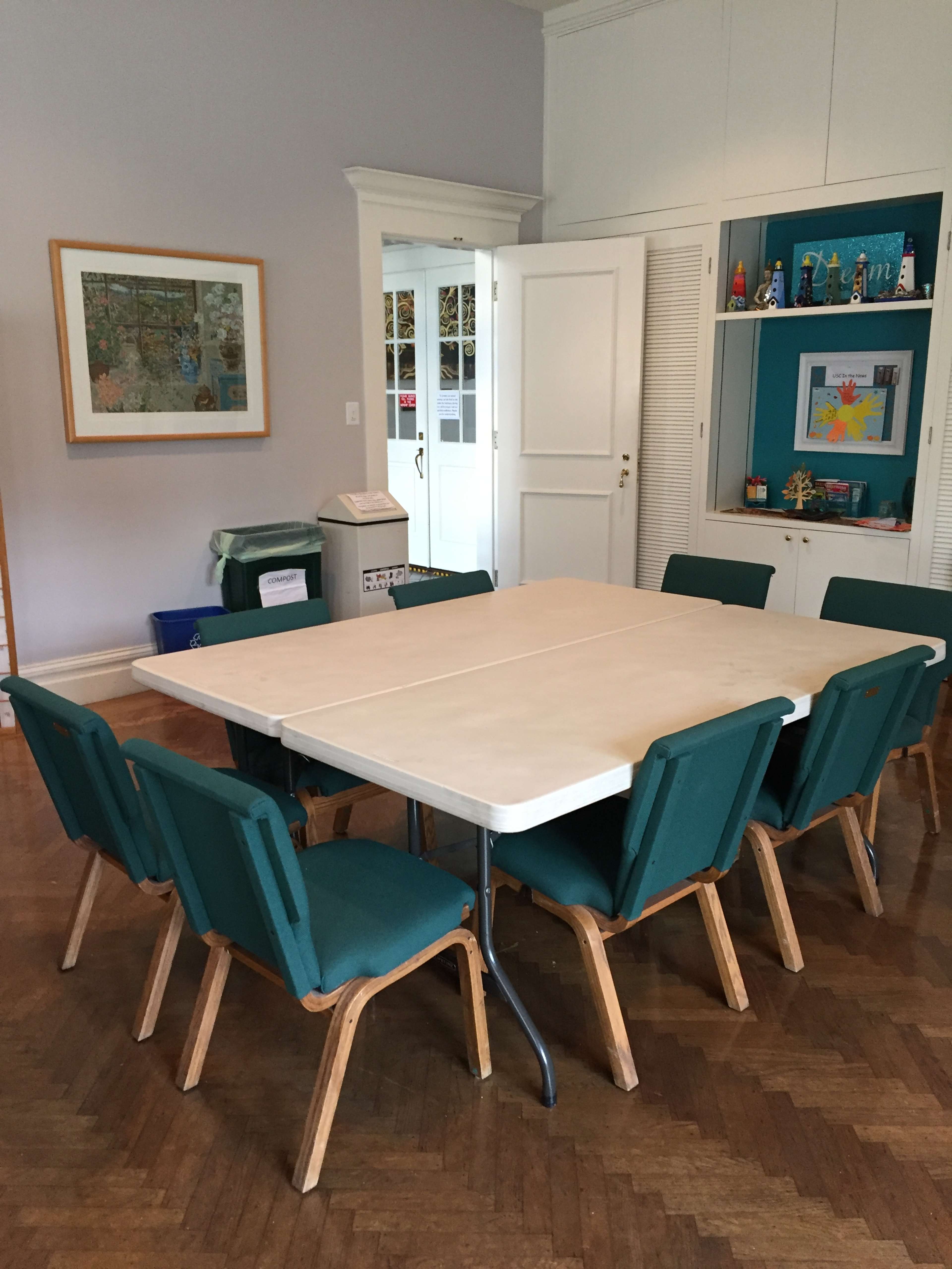 A rectangular table surrounded by eight teal chairs is set up in a room with light-colored walls and a framed artwork on display.