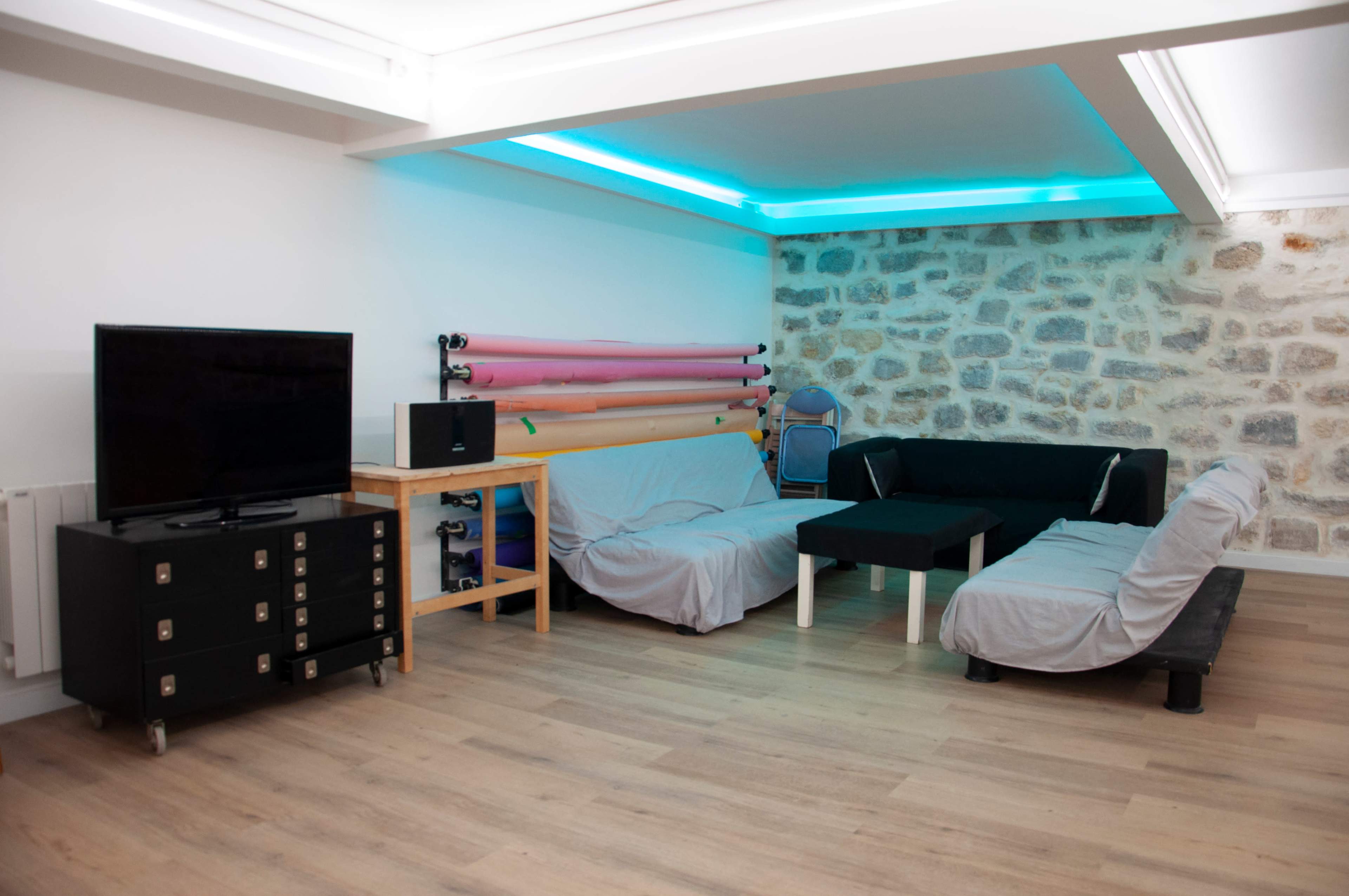 A modern living space with two couches covered in light fabric, a black coffee table, a television on a rolling cabinet, and colorful rolls of material stored against a stone wall.
