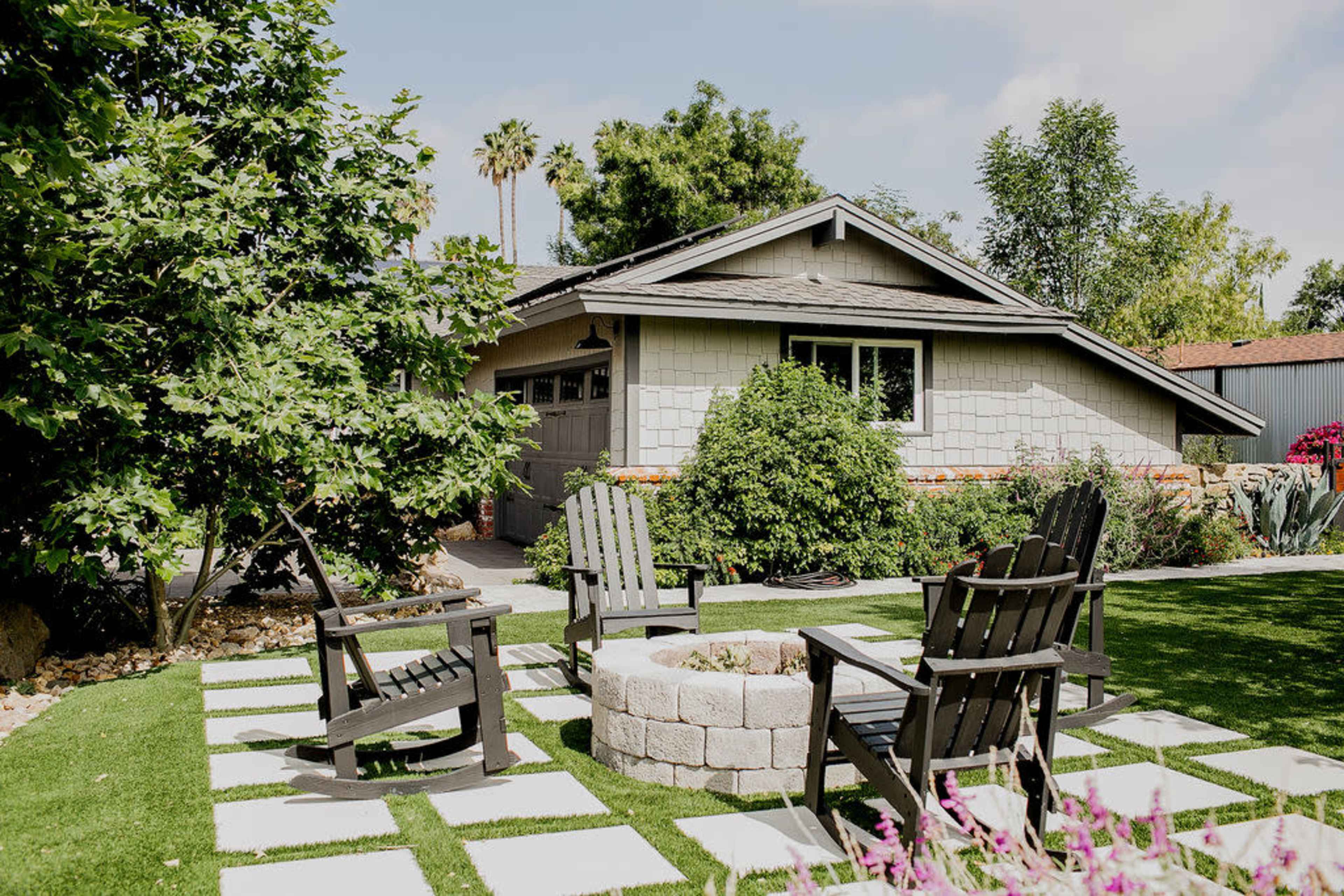 A backyard patio features four wooden chairs arranged around a circular fire pit, with a house and trees visible in the background.