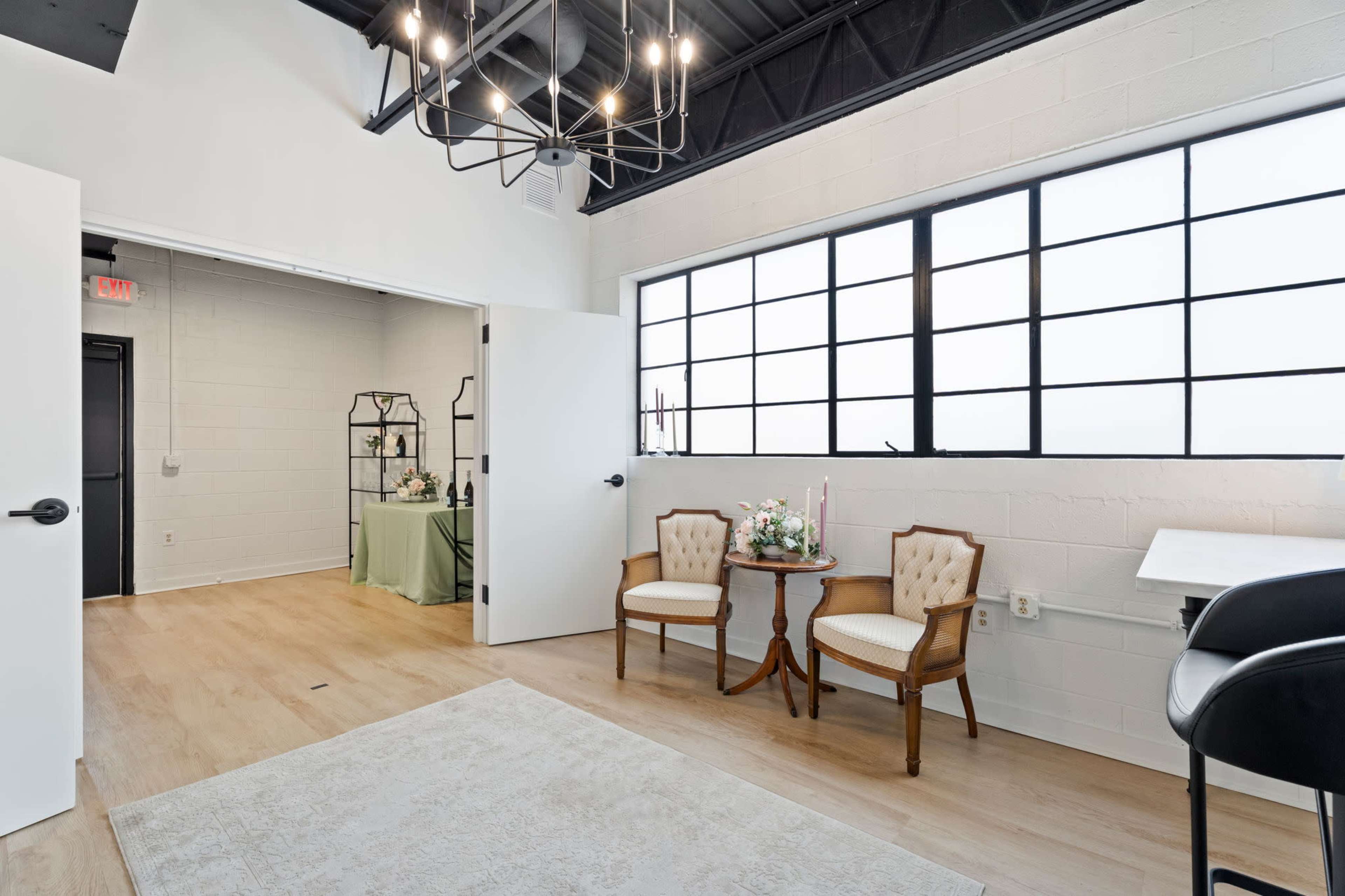 The image shows a bright, modern room featuring two chairs and a small table near a large window, with light-colored walls and wooden flooring.