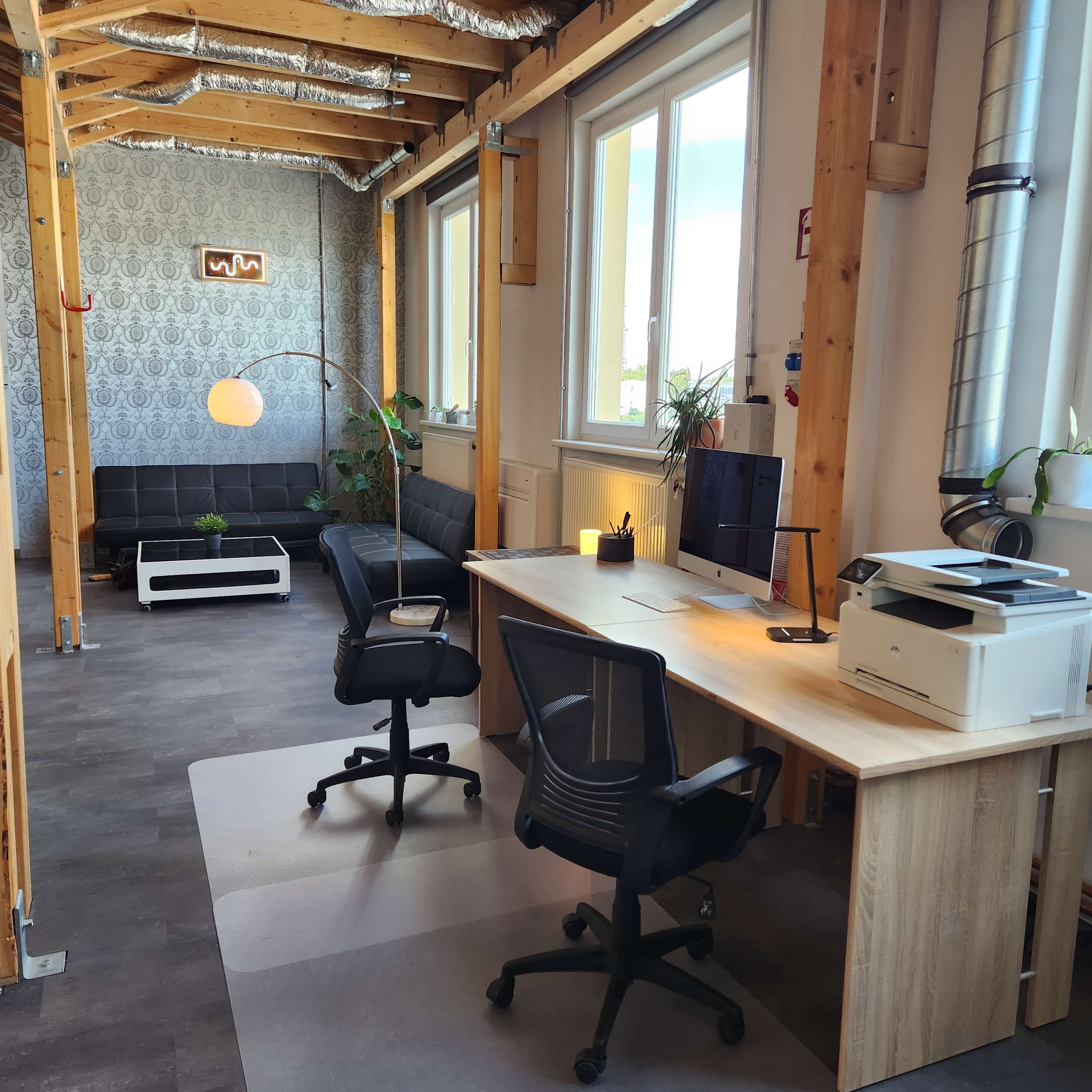 The image shows a modern office space with a wooden desk, two black ergonomic chairs, a computer setup, a printer, and a couch in a well-lit environment with plants.