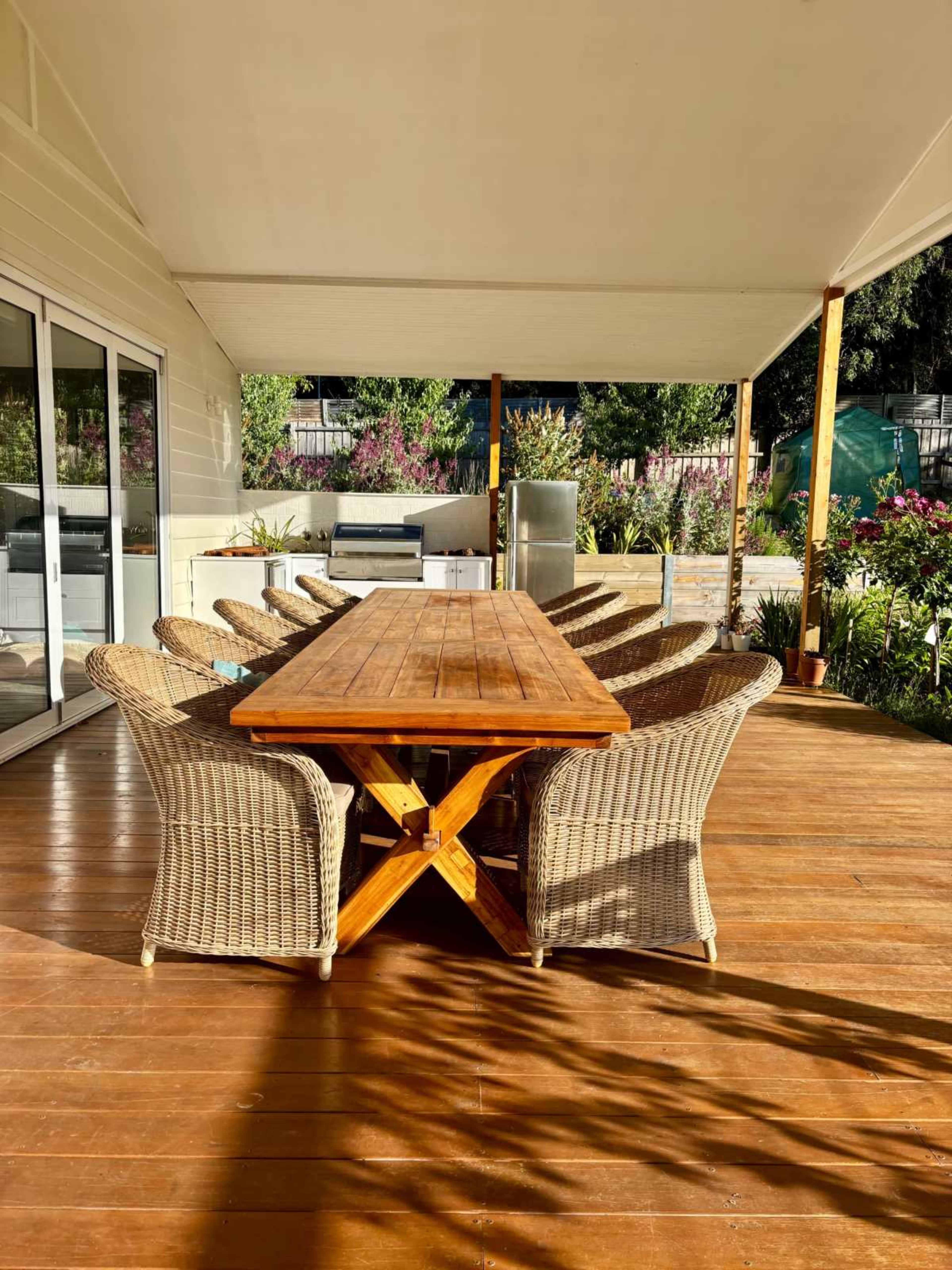 A wooden dining table surrounded by wicker chairs is positioned on a deck under a covered patio, with greenery visible in the background.