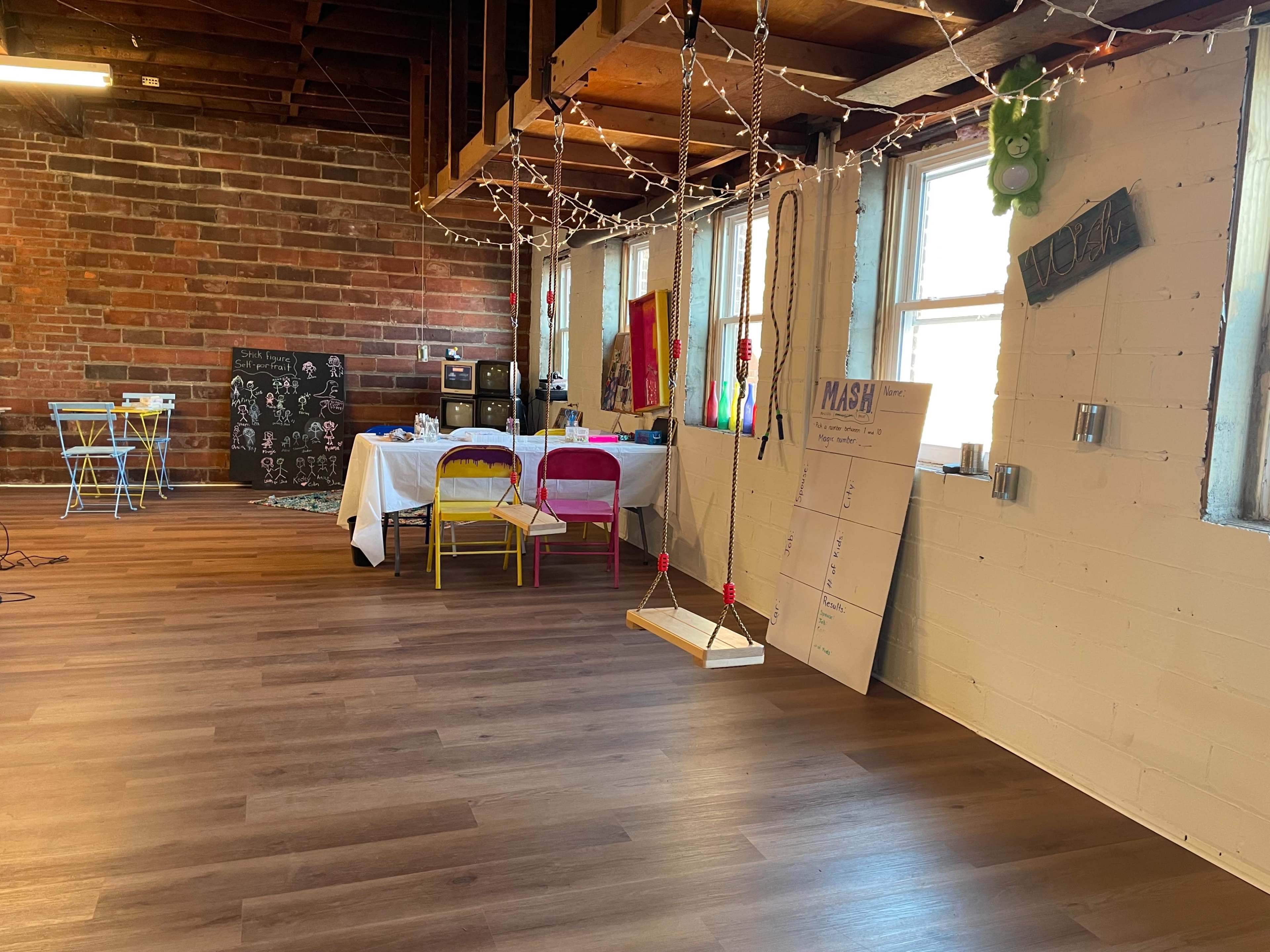 A spacious room with wooden floors, illuminated by string lights, featuring a swing, colorful chairs, and a table set with supplies against a brick wall.
