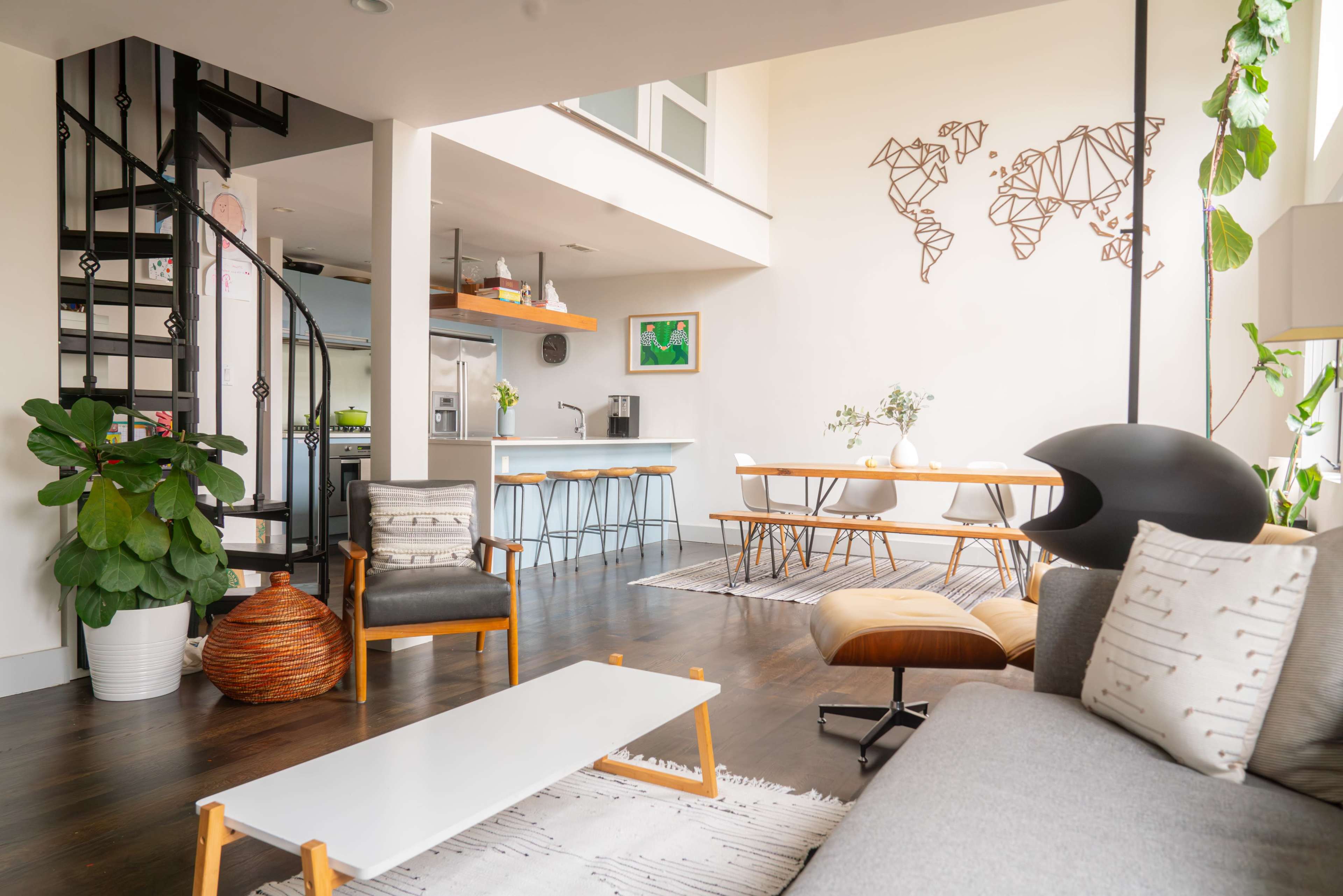 The image shows a modern, open-concept living and dining area featuring a spiral staircase, wooden dining table, and plants.