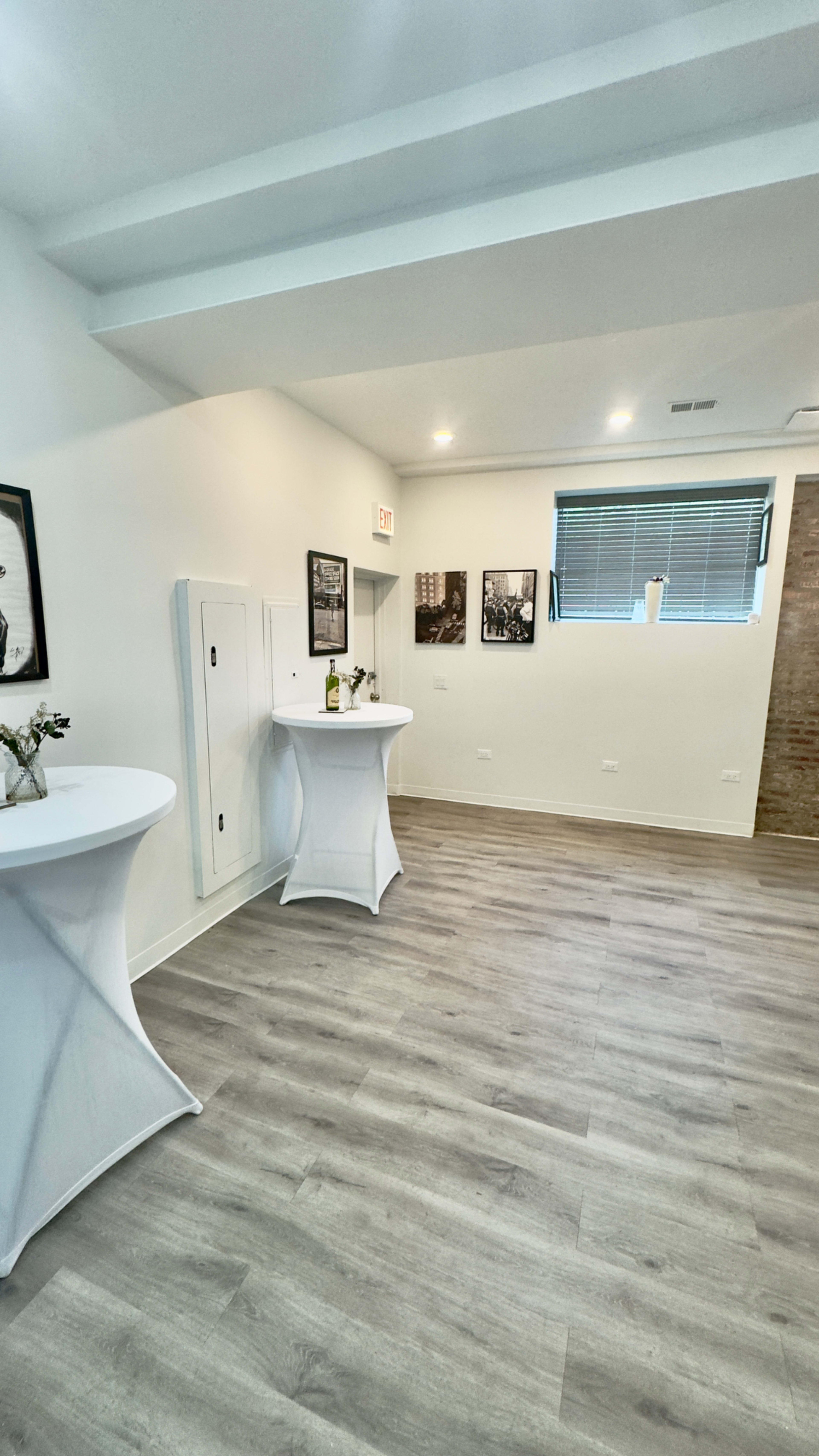 The image shows a spacious, bright room with light wood flooring, featuring two tall, white decorative tables and photographs on the walls.