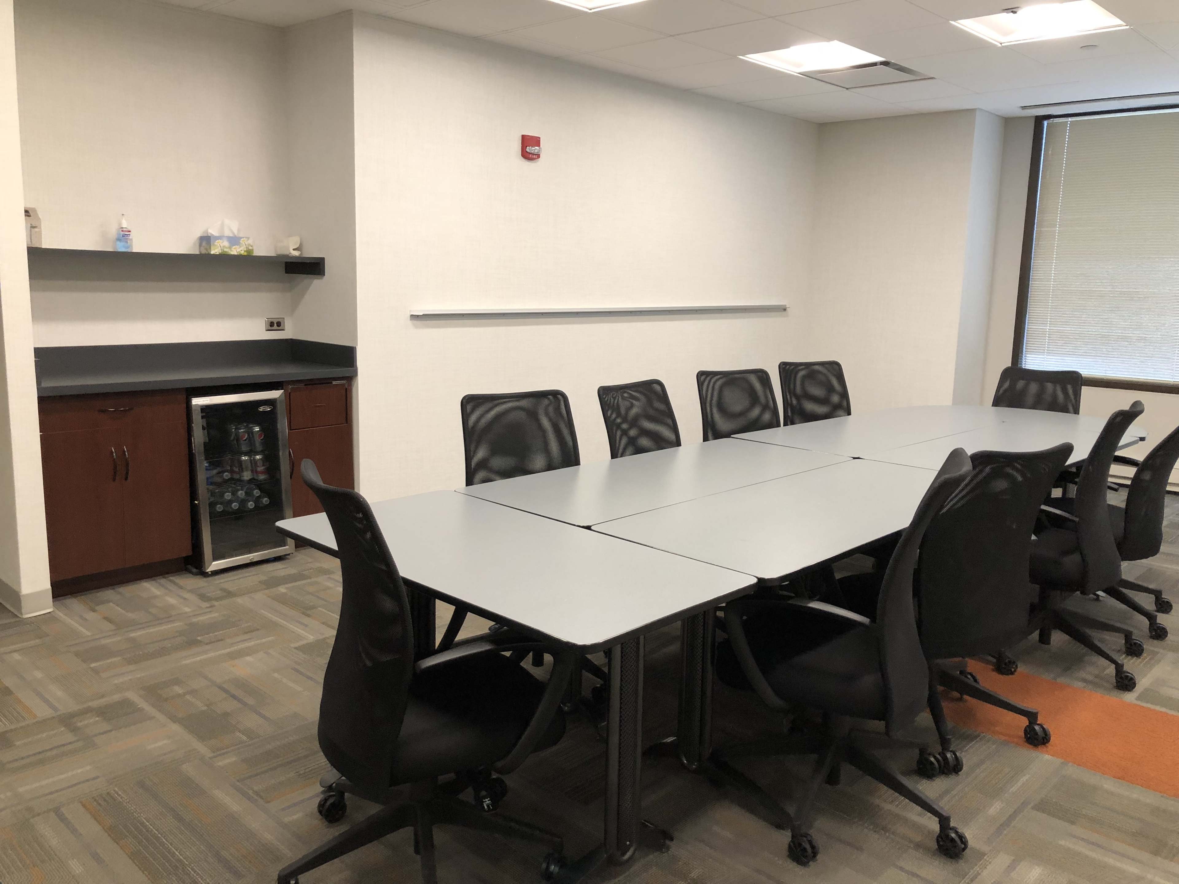 The image shows a conference room with a large table and black rolling chairs, a small kitchenette with a fridge, and a window providing natural light.