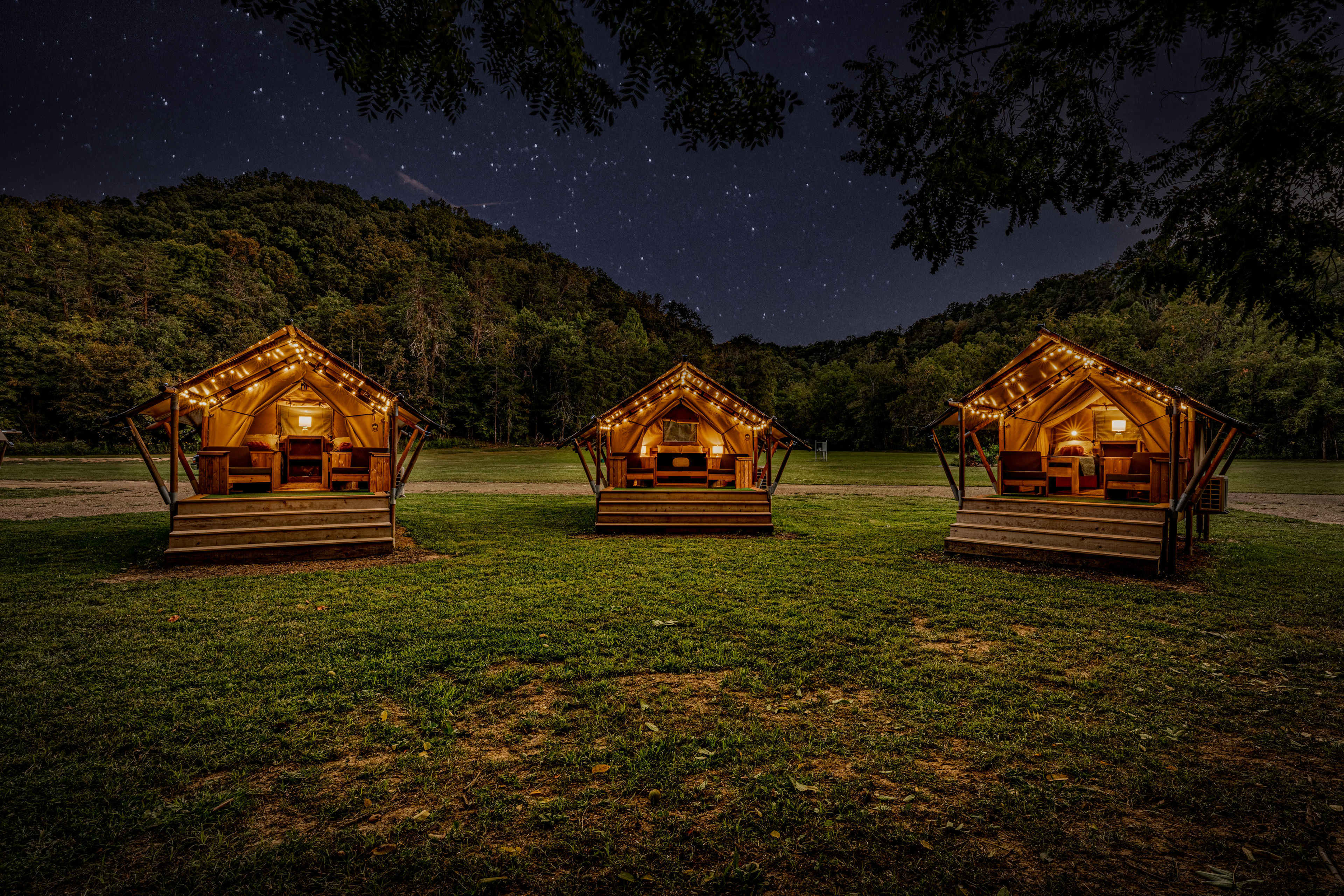 Three illuminated glamping tents are set on a grassy area under a starry night sky, surrounded by wooded hills.