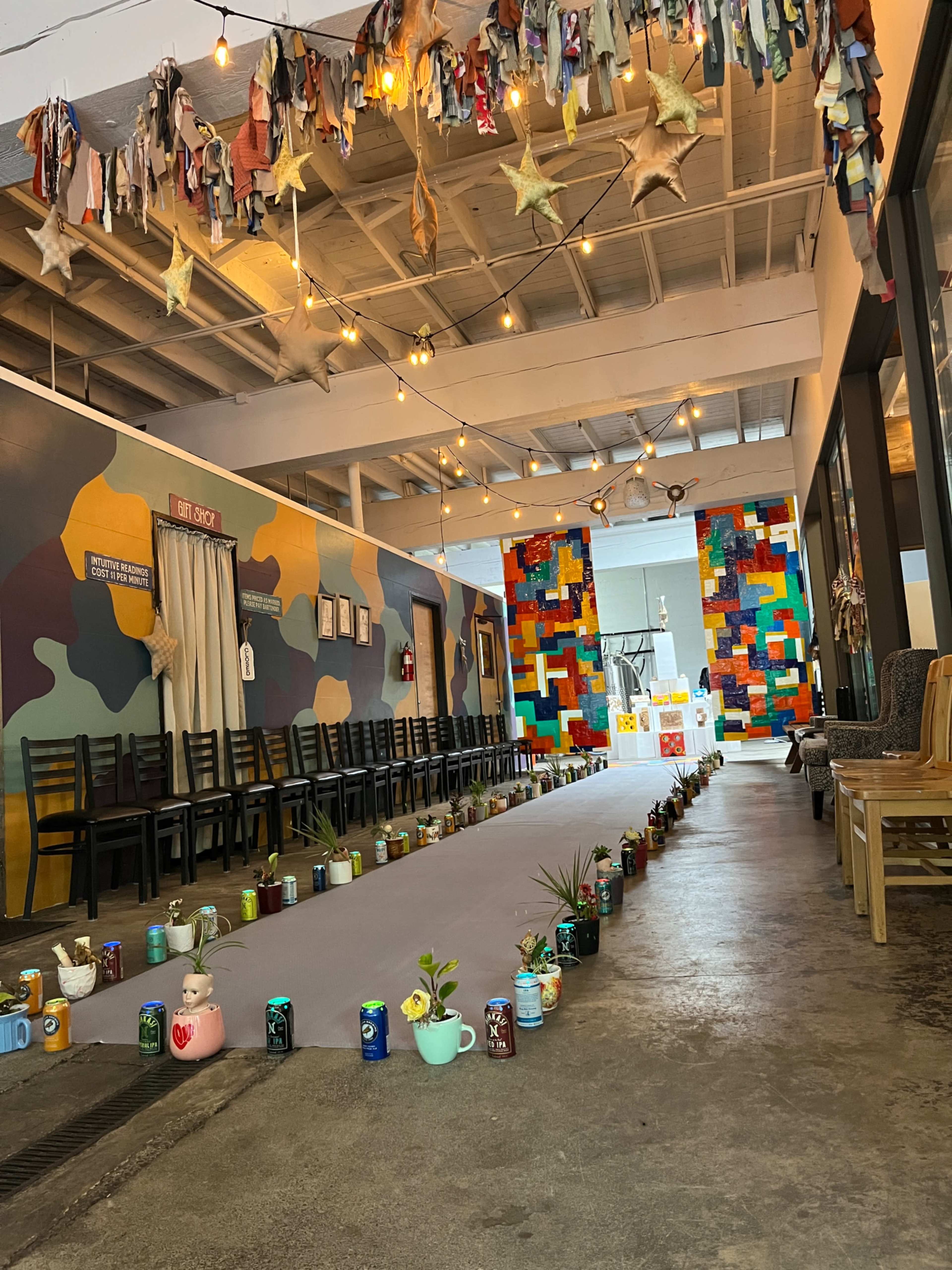 The image shows a colorful indoor space with a long walkway bordered by various decorative cans and plants, leading towards a vibrant mural at the far end.