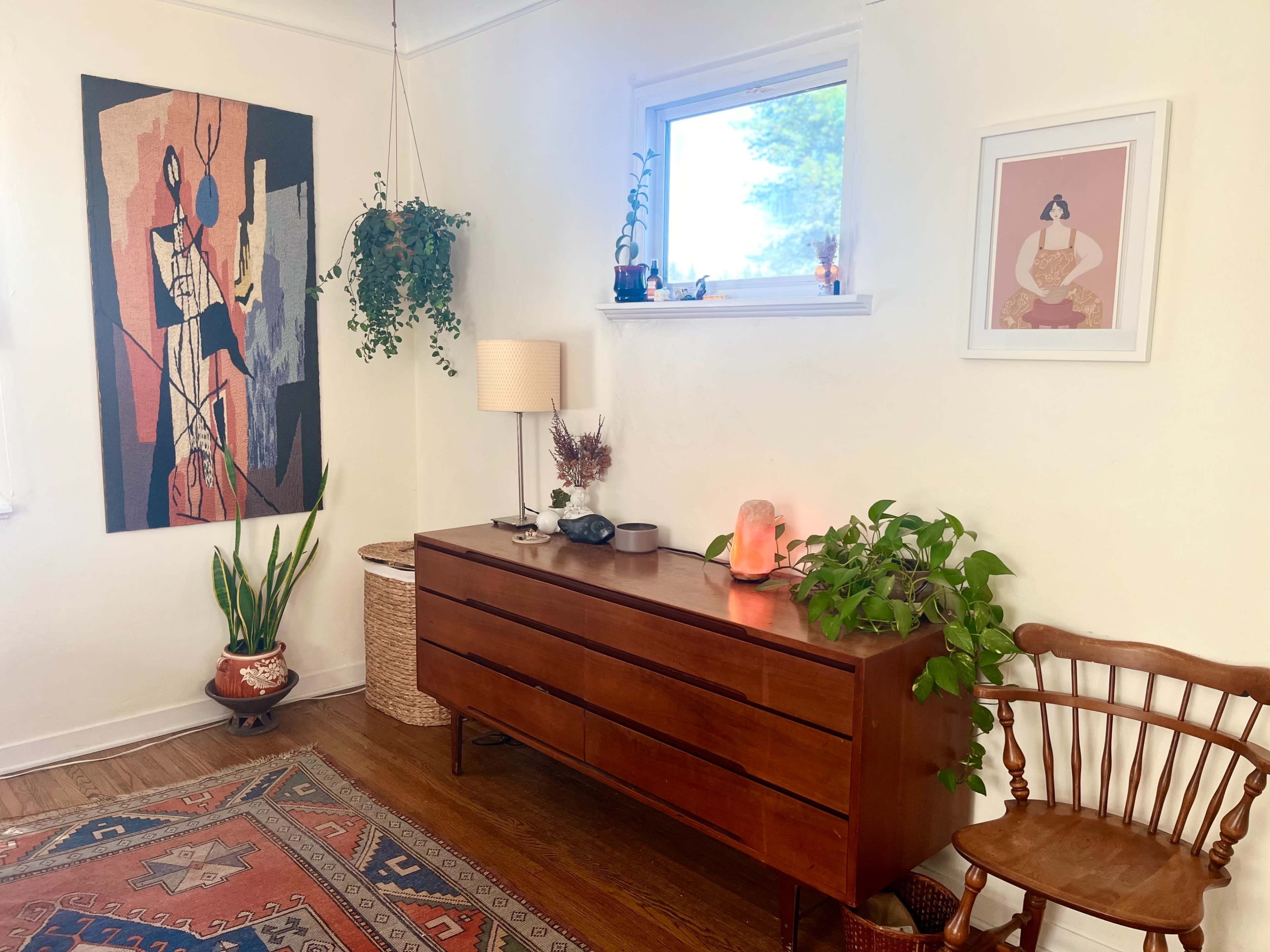 The scene features a cozy room with a wooden dresser, a potted plant, wall art, and a lamp, complemented by a woven basket and a chair.