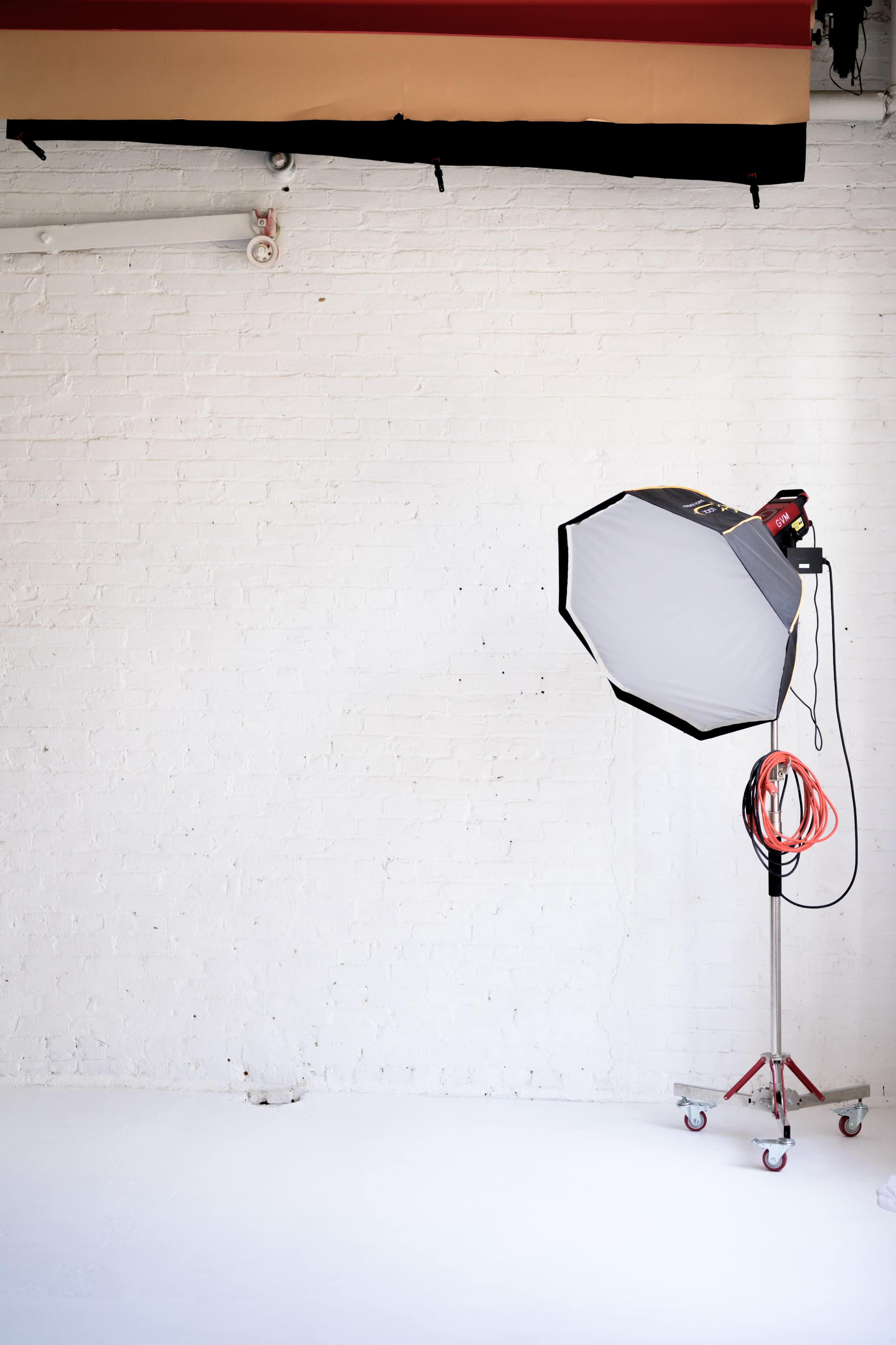 A photography studio is depicted with a large softbox light on a stand against a white brick wall.