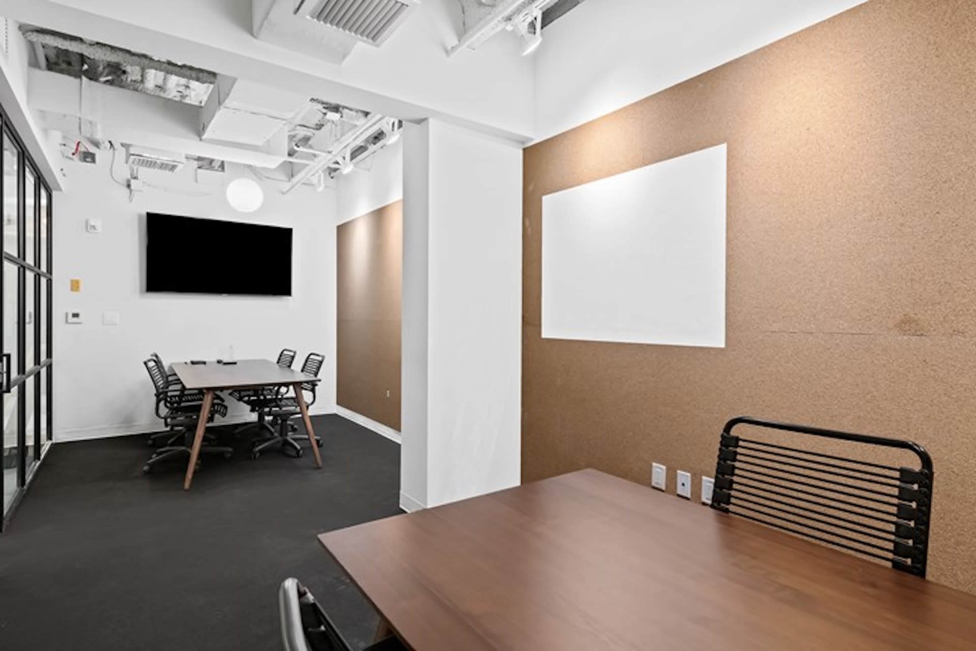 A conference room features a long wooden table, black chairs, and cork walls, with a television mounted on one side.
